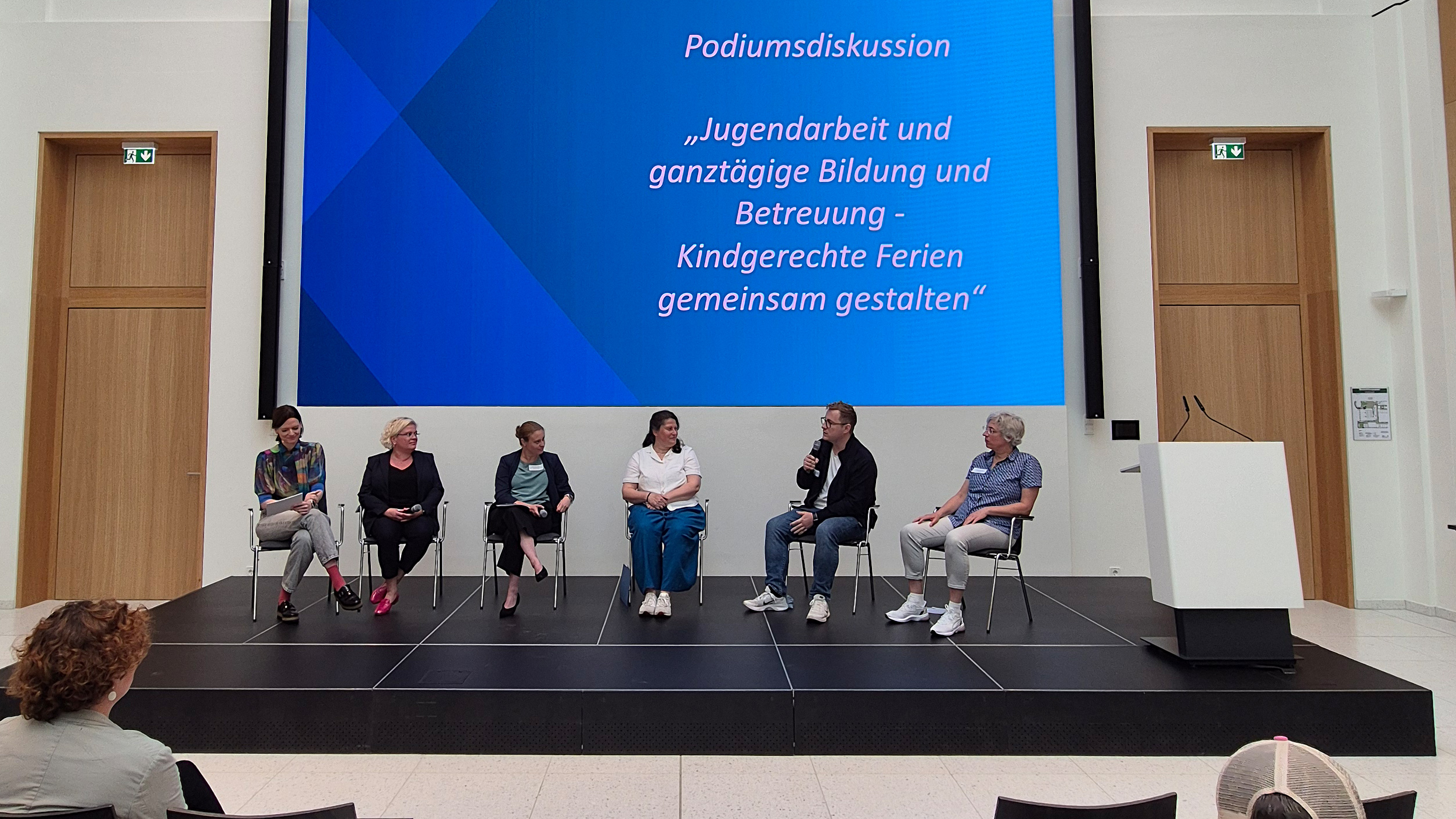 Podiumsdiskussion mit sechs Personen auf Stühlen vor großem Bildschirm mit Text zu Jugendarbeit und ganztägiger Bildung und Betreuung.
