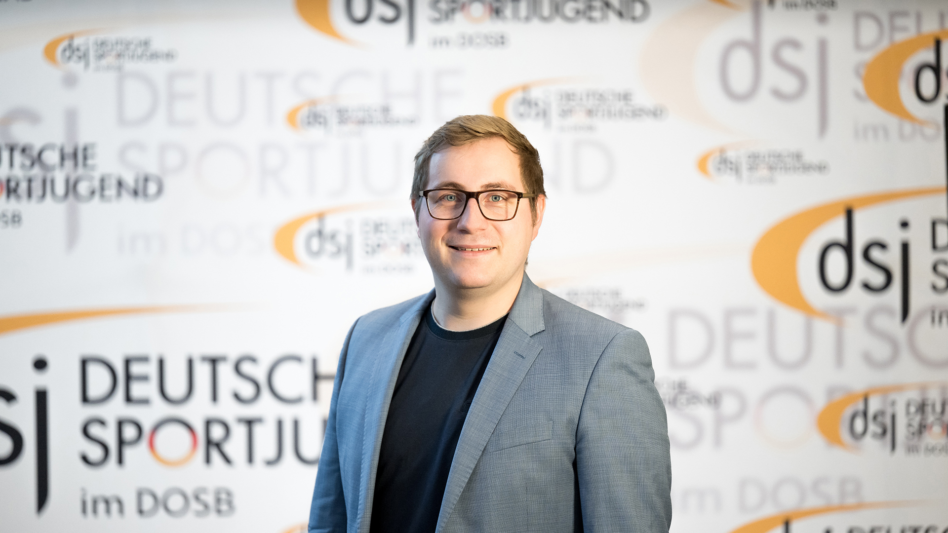 Julian Lagemann steht vor einer Fotowand der Deutschen Sportjugend (dsj) im DOSB und trägt ein graues Sakko über einem dunklen Shirt