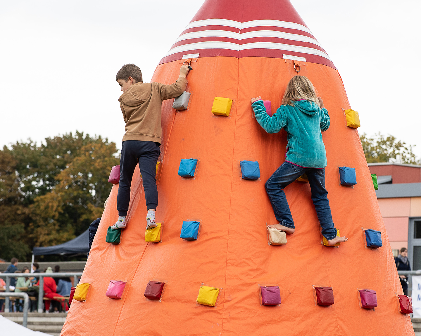 Zwei Kinder klettern an einer bunten Kletterwand in Form eines Kegels. Das eine Kind trägt einen braunen Pullover und das andere ein grünes Sweatshirt. Im Hintergrund sind Menschen und Bäume zu sehen.