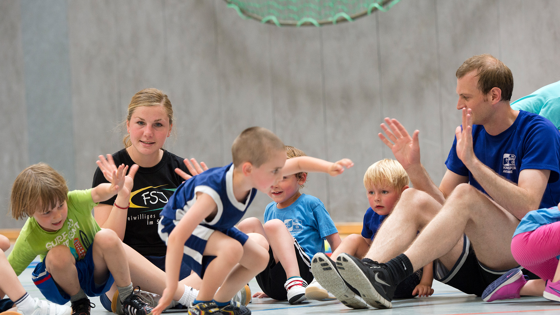 Mehrere Kinder und ein Erwachsener sitzen auf dem Boden in einem Kreis. Die Kinder tragen sportliche Kleidung in verschiedenen Farben, darunter grün, blau und schwarz. Der Erwachsene trägt ein blaues T-Shirt und schwarze Shorts. Einige der Kinder und der Erwachsene heben die Hände, als würden sie sich abklatschen oder eine spielerische Geste machen. Der Hintergrund zeigt einen hellen Raum mit einem hellblauen Sitzkissen oder Polster an der Wand.