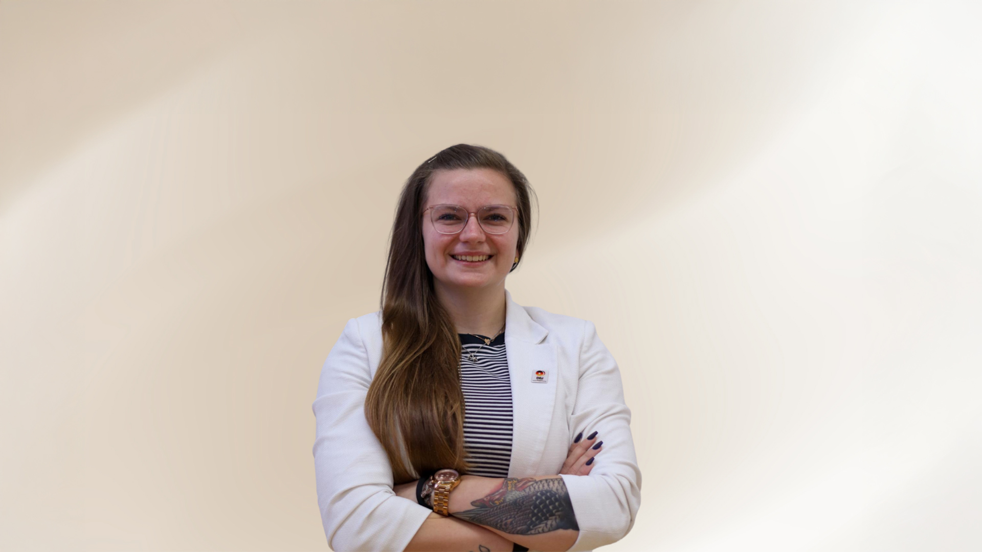 Eine Frau, Lili Oesten steht vor hellem Hintergrund mit verschränkten Armen, trägt einen hellen Blazer über einem gestreiften Oberteil und blickt frontal in die Kamera.