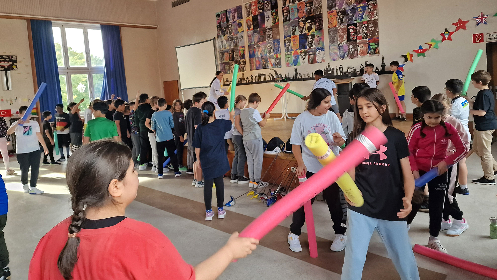 In einem großen, hellen Raum mit hohen Fenstern und einer Wand, die mit bunten Postern und Sternen dekoriert ist, stehen viele Kinder und Jugendliche verteilt. Sie halten bunte Schaumstoffstäbe in verschiedenen Farben wie Rot, Gelb, Blau und Grün. Die Kinder sind in Bewegung, einige scheinen sich gegenseitig mit den Stäben zu berühren oder zu spielen. Im Hintergrund ist eine weiße Leinwand an der Wand zu sehen. Die Atmosphäre wirkt lebendig und aktiv, passend zum Kontext des Zukunftslabors der Fechtjugend, das Bewegung fördert.
