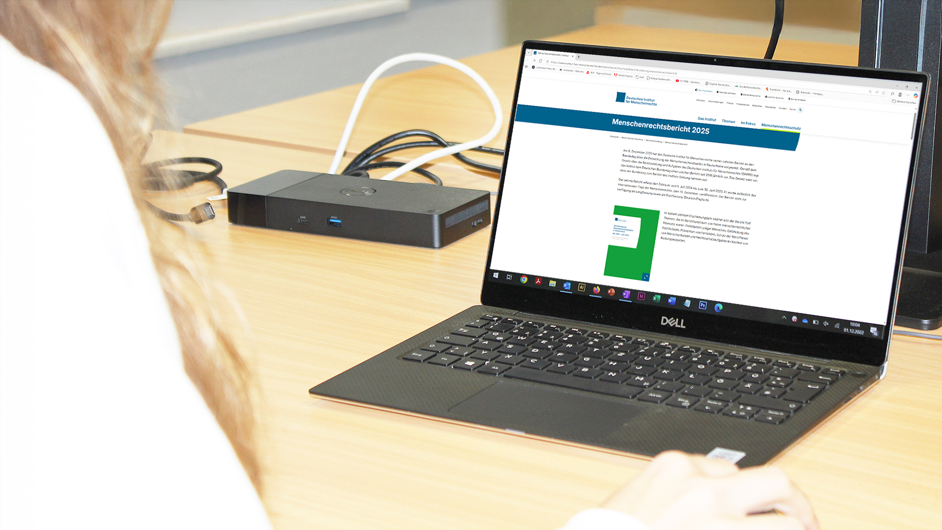 Eine Person mit langen, blonden Haaren sitzt an einem hellen Holztisch und arbeitet an einem schwarzen Dell-Laptop. Auf dem Bildschirm ist eine Webseite mit dem Titel 'Menschenrechtsbericht 202' zu sehen, darunter ein grünes Buchcover. Neben dem Laptop liegt ein schwarzes elektronisches Gerät mit mehreren Kabeln, die zum Laptop führen.