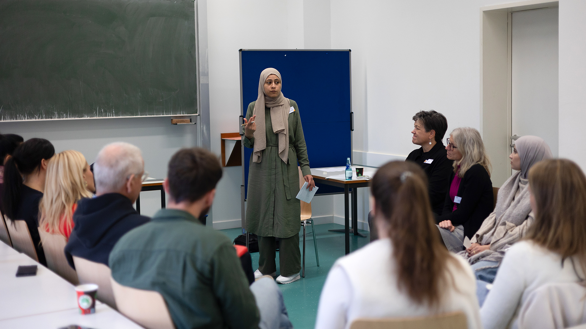 In einem hellen Seminarraum steht eine Frau mit einem hellen Kopftuch und einem langen, olivgrünen Kleid vor einer Gruppe sitzender Personen. Sie hält ein Blatt Papier in der linken Hand und gestikuliert mit der rechten Hand. Die Zuhörenden sitzen in einem Halbkreis auf weißen Stühlen und blicken zur Sprecherin. Im Hintergrund ist eine grüne Tafel an der Wand sowie eine blaue Stellwand zu sehen. Auf einem Tisch hinter der Sprecherin steht eine Wasserflasche. Die Szene zeigt eine Diskussions- oder Vortragssituation während der Abschlusstagung des Netzwerks „Antirassismus im organisierten Sport (NAniS)“.