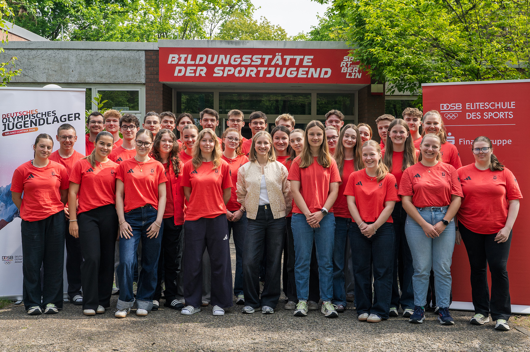 Eine Gruppe von 30 jungen Erwachsenen in roten T-Shirts steht vor einem Gebäude mit dem Schriftzug "Bildungsstätte der Sportjugend". Sie lächeln und präsentieren sich gemeinsam, während zwei Banner auf beiden Seiten Informationen zur Sportjugend zeigen.