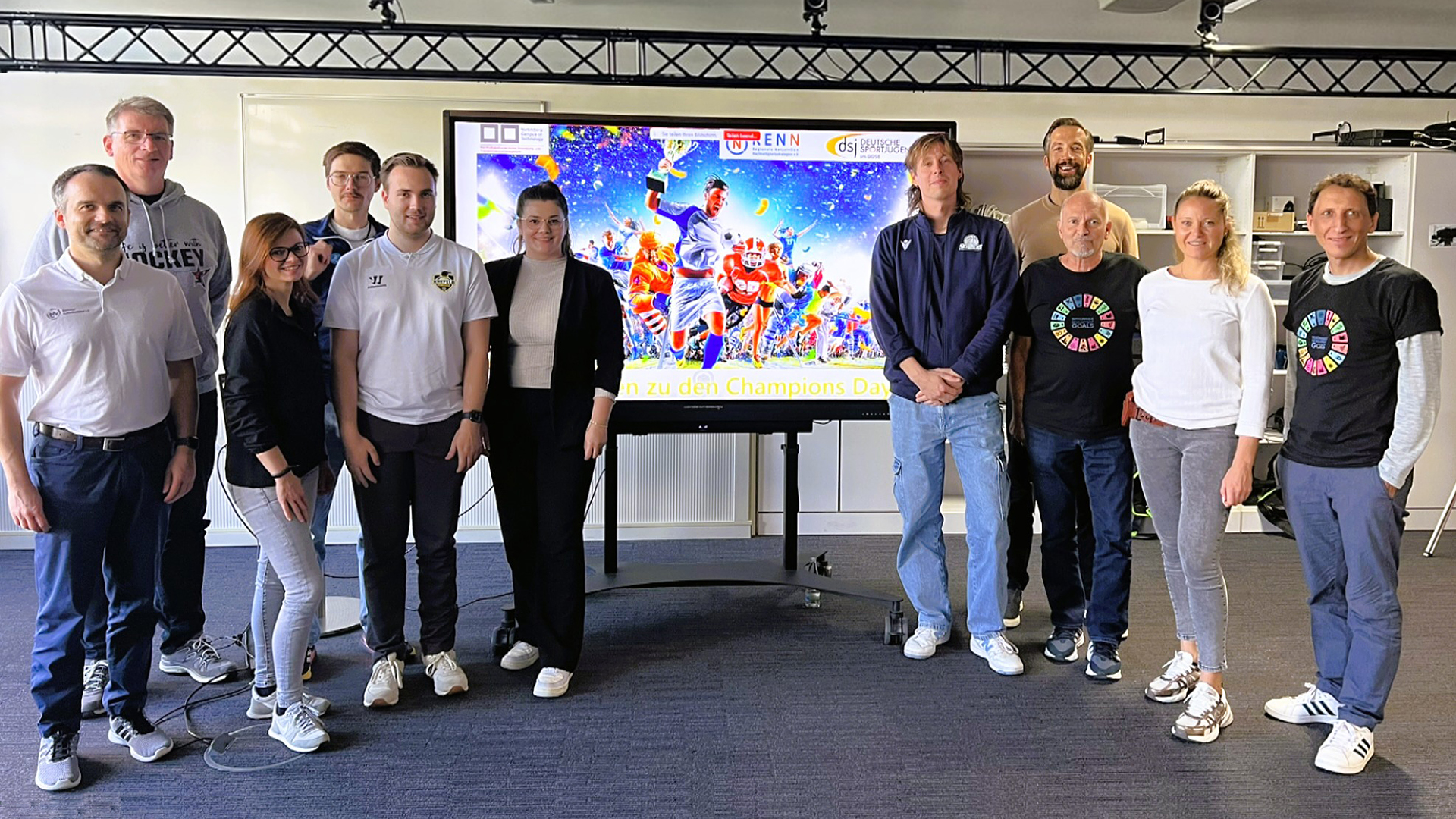 Gruppe von Personen und frischgebackene Nachhaltigkeitsmanager*innen steht vor einem großen Bildschirm mit bunter Sportgrafik und Text 'zu den Champions Day' in einem Veranstaltungsraum.