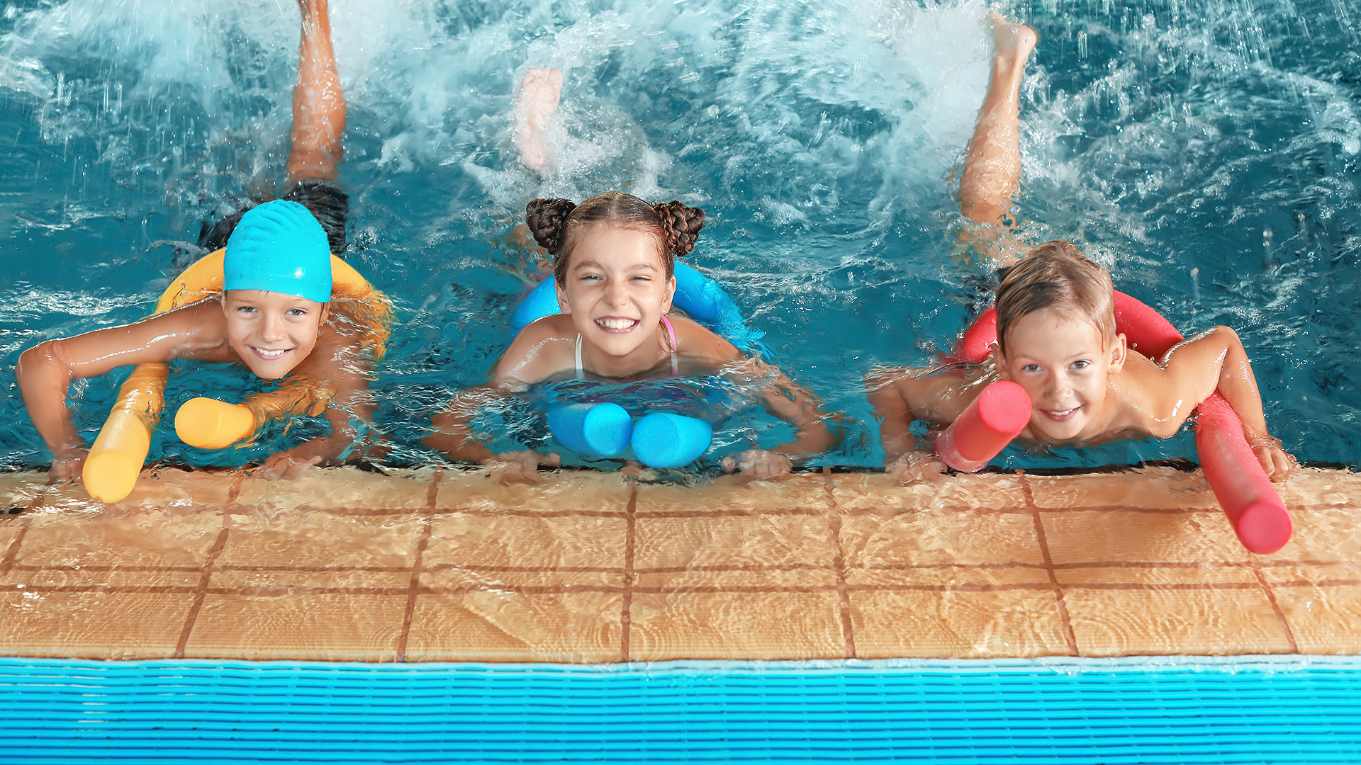 Drei Kinder liegen bäuchlings am Beckenrand eines Schwimmbeckens und paddeln mit bunten Schwimmnudeln im Wasser.