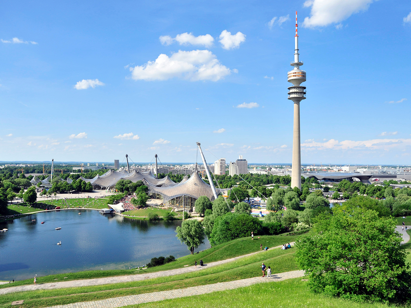 Blick auf den Olympiapark in München mit dem Olympiastadion und dem Olympiaturm. Im Vordergrund ein See und grüne Wiesen, im Hintergrund eine klare blaue Himmel mit einigen Wolken.