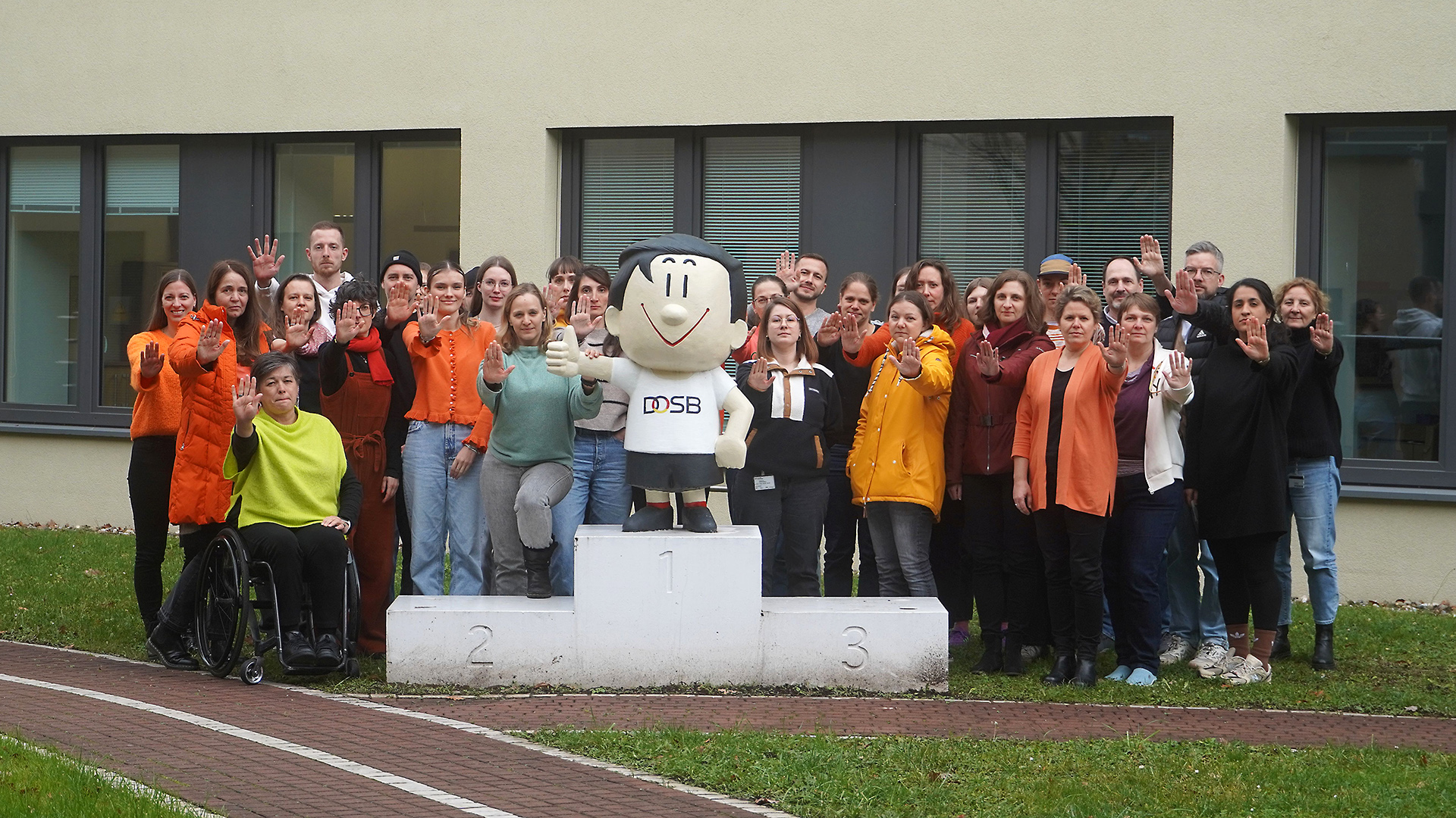Eine Gruppe von Menschen steht vor einem Gebäude mit mehreren Fenstern. In der Mitte steht eine weiße, maskottchenartige Figur mit dem Schriftzug 'DOSB' auf der Brust auf einem dreistufigen Podest. Die Personen tragen überwiegend orangefarbene Kleidung und heben die rechte Hand mit ausgestreckten Fingern, was eine Geste des Stopps symbolisiert. Eine Person im Rollstuhl ist vorne links zu sehen. Der Hintergrund zeigt eine Wiese und einen Fußweg vor dem Gebäude.