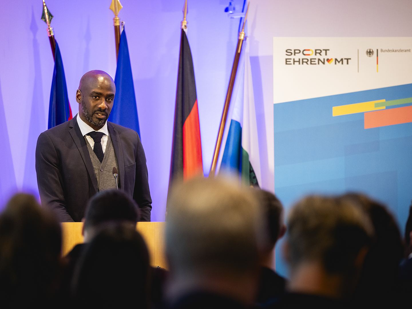 Otto Addo steht an einem Rednerpult und spricht. Im Hintergrund sind mehrere Flaggen zu sehen. Rechts ist ein Plakat mit dem Schriftzug "SPORT EHRENAMT" und dem Logo des Bundesministeriums. 