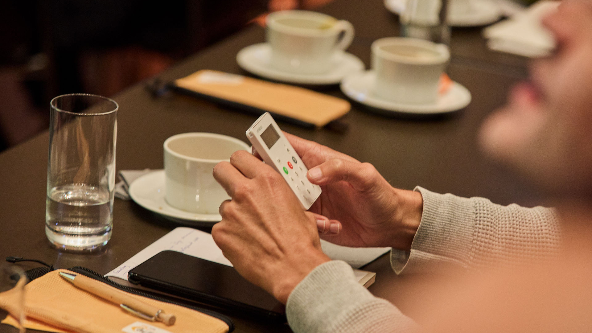 Hände einer Person halten ein kleines weißes Abstimmungsgerät mit mehreren farbigen Tasten umd für die Resolution deim dsj-Hauptausschuss "Für eine resiliente Gesellschaft - Freiwilligendienste im Sport stärken!" abzustimmen. Im Hintergrund stehen auf einem Tisch zwei weiße Kaffeetassen auf Untertassen, ein Glas mit Wasser und ein braunes Notizbuch. Die Person trägt einen hellgrauen Pullover, der Arm ist leicht angewinkelt.