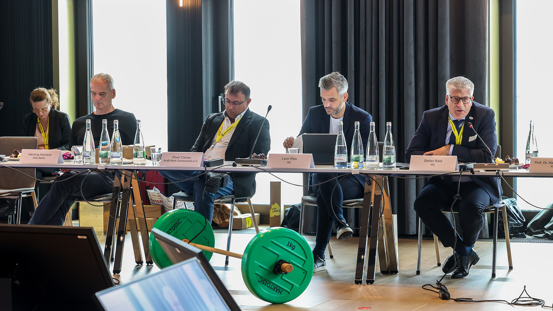 Personen sitzen an einem Konferenztisch mit Mikrofonen, Wasserflaschen und Namensschildern, im Hintergrund Fenster mit Vorhängen.