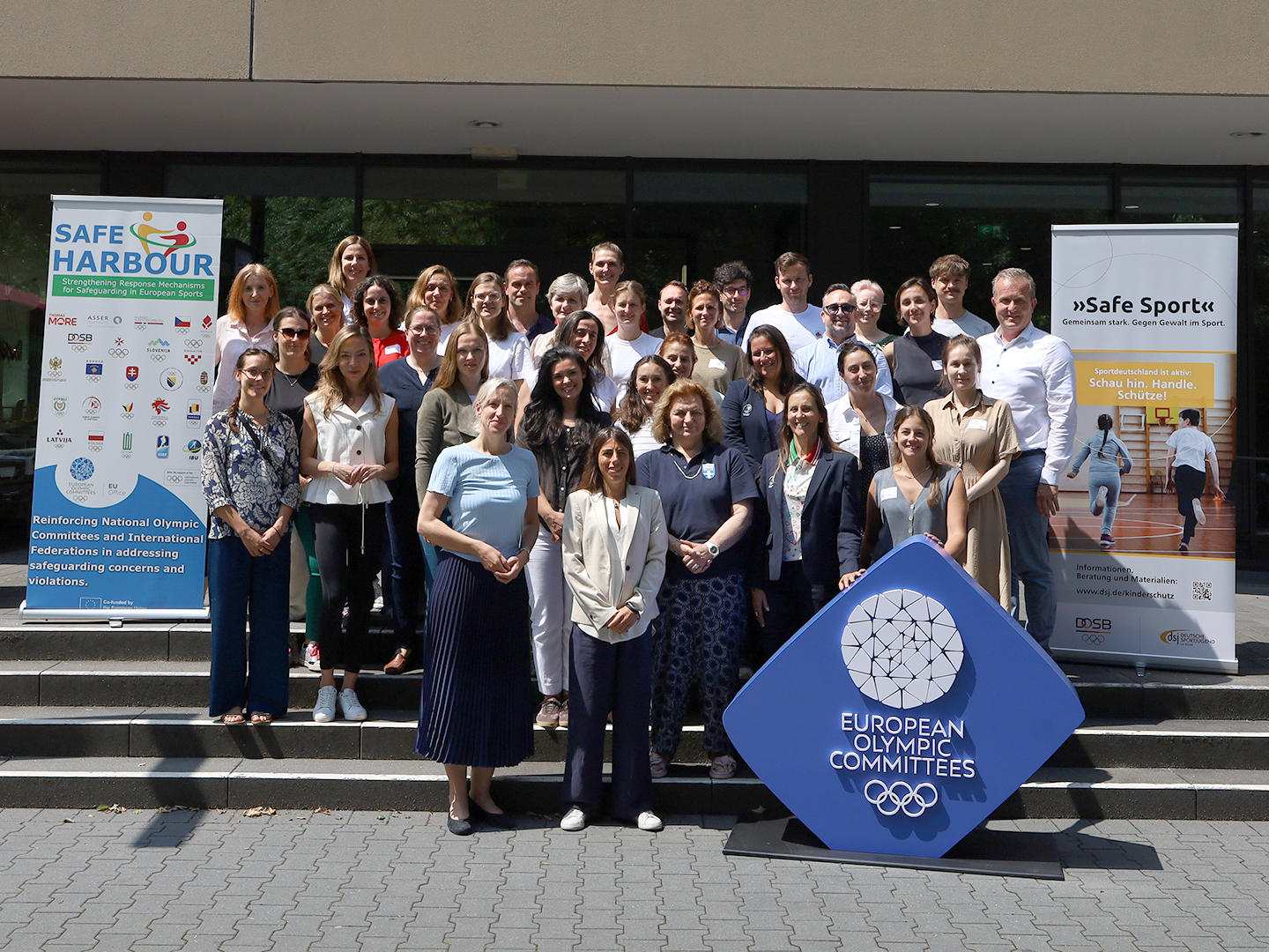 Eine Gruppe von etwa 40 Personen steht vor einem Gebäude, umgeben von Bannern, die mit olympischen Themen und Sportorganisationen zu tun haben. Im Vordergrund befindet sich ein großes, blaues Logo der Europäischen Olympischen Komitees. Die Teilnehmer lächeln und posieren für das Foto.