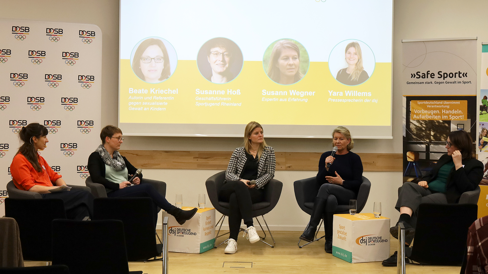 Fünf Frauen sitzen in einem Halbkreis auf Stühlen und sprechen miteinander. Im Hintergrund ist eine Leinwand mit einer Präsentation zu sehen, auf der vier runde Porträtbilder mit den Namen Beate Kriechel, Susanne Hoß, Susann Wegner und Yara Willems zu erkennen sind. Rechts steht ein Banner mit dem Schriftzug »Safe Sport« und dem Text »Gemeinsam stark. Gegen Gewalt im Sport. Vorbeugen. Handeln. Aufarbeiten im Sport«. Die Frauen tragen unterschiedliche Kleidung, darunter ein rotes Oberteil, ein kariertes Jackett und dunkle Hosen. Die Szene wirkt wie eine Podiumsdiskussion.
