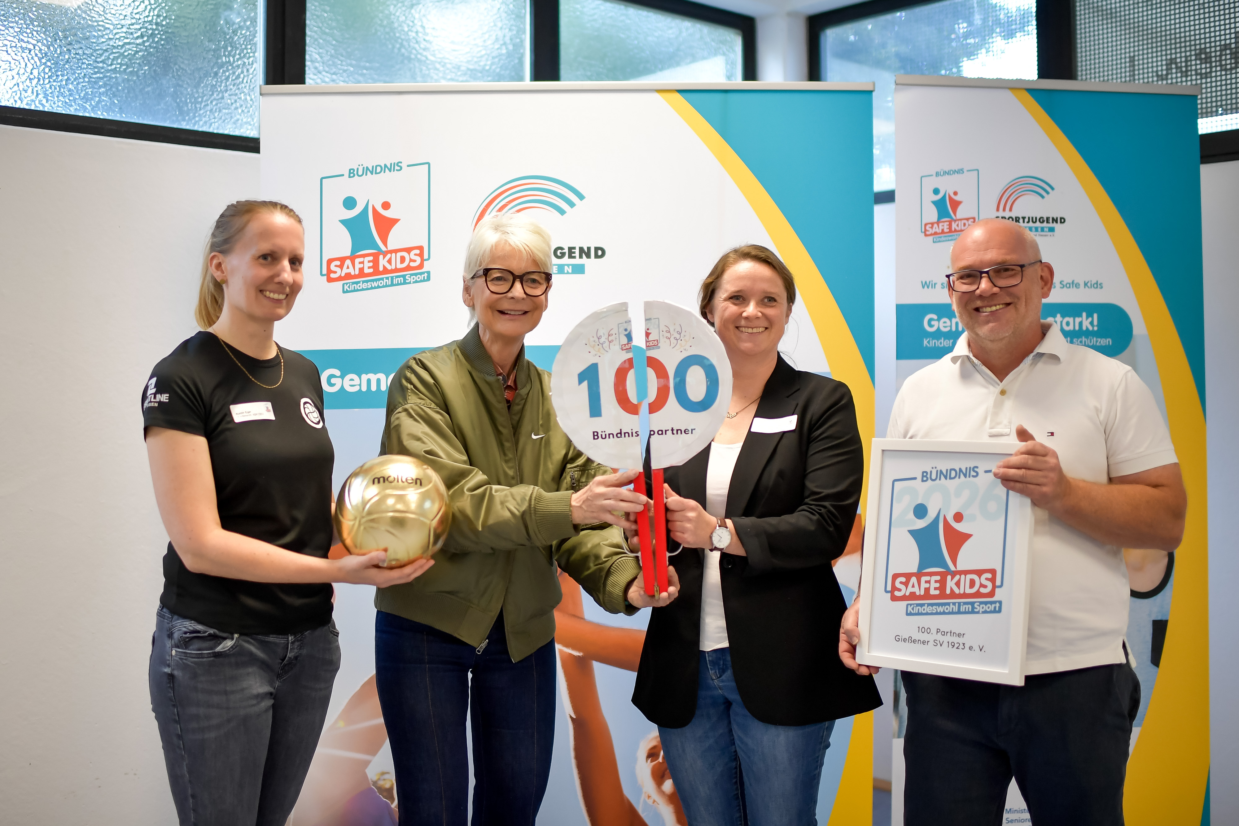 Vier Personen stehen zusammen und halten verschiedene Objekte in den Händen, darunter eine goldene Kugel und ein Schild mit der Zahl 100. Im Hintergrund sind Banner mit dem Logo von "Bündnis Safe Kids" zu sehen. Die Atmosphäre wirkt feierlich und engagiert.