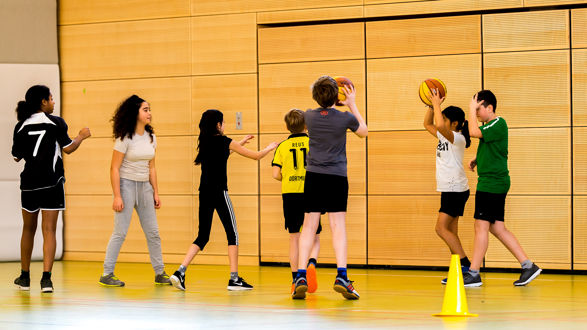 Kinder stehen in einer Sporthalle und führen gemeinsam Übungen mit Bällen aus. Die Szene zeigt aktiven Schulsport und gemeinsames Bewegen im Unterricht.