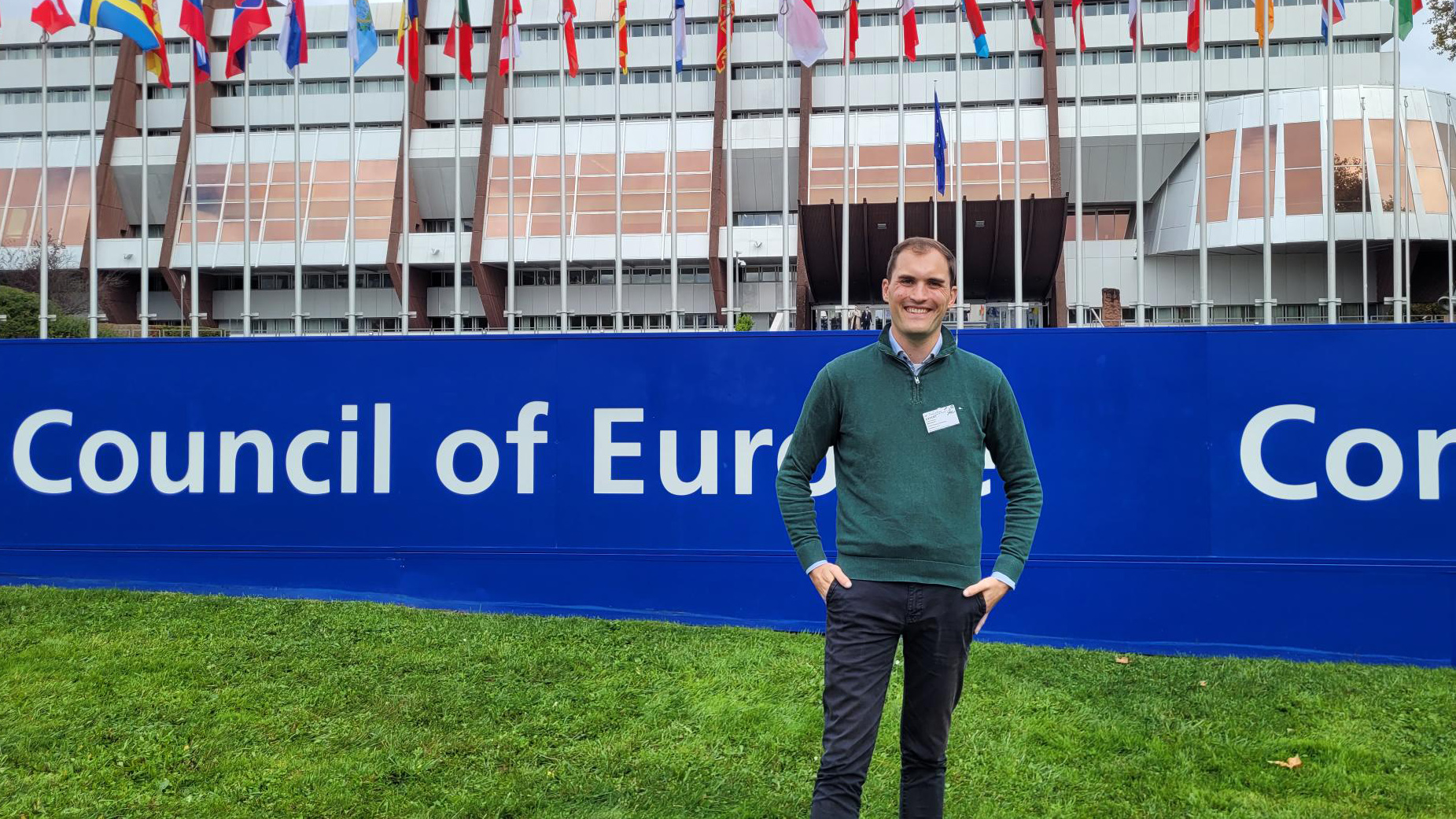 Ein Mann, Luca Wernert Vice-Chair von ENGSO Youth und dsj-Vorstandsmitglied steht auf einer Rasenfläche vor einem blauen Banner mit der weißen Aufschrift 'Council of Europe'. Er trägt einen grünen Pullover über einem hellblauen Hemd, eine schwarze Hose und hat die Hände in den Hosentaschen. Im Hintergrund sind zahlreiche Flaggen verschiedener Länder vor einem mehrstöckigen Gebäude zu sehen.