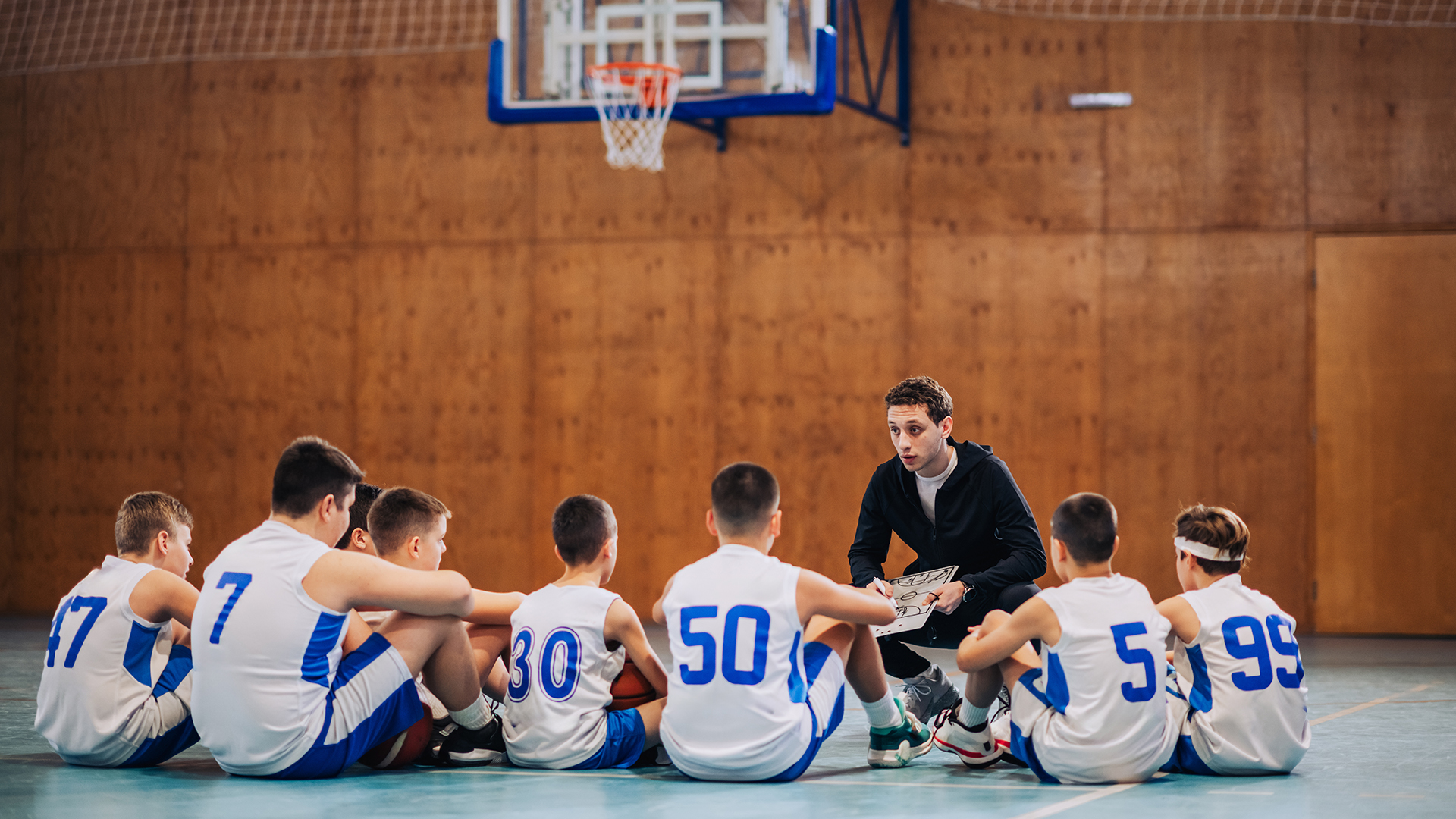Basketballtrainer spricht in einer Sporthalle mit seiner Jugendmannschaft, die im Kreis sitzt – symbolisches Motiv für Teamgeist, Förderung junger Talente und den politischen Rückenwind für Bewegung, Teilhabe und die deutsche Bewerbung für Olympische und Paralympische Spiele.