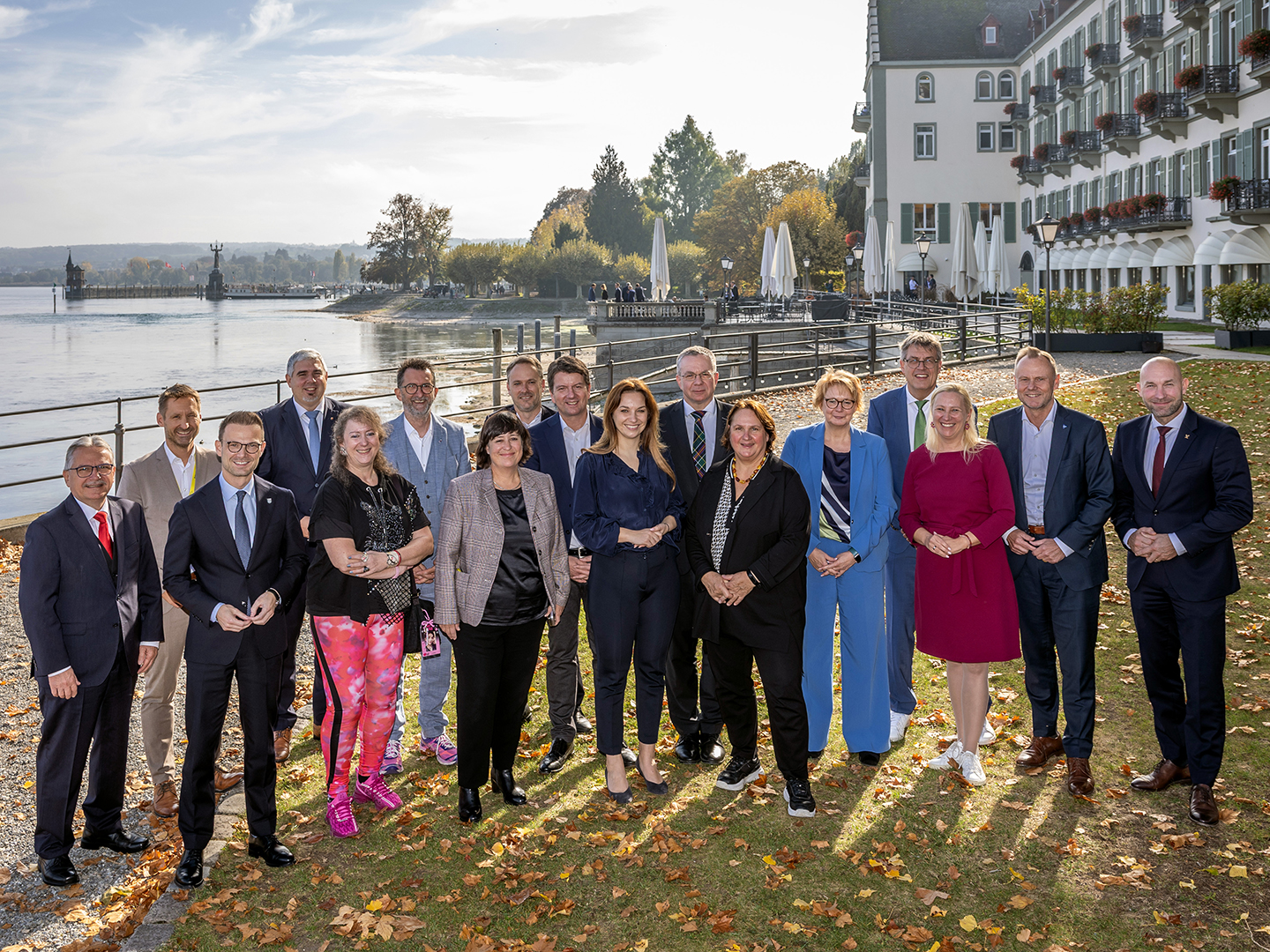 Eine Gruppe von 20 Personen steht am Ufer eines Sees, umgeben von Bäumen und einem Hotel. Es ist ein sonniger Tag im Herbst, und die Menschen lächeln in die Kamera. Einige tragen formelle Kleidung, während andere lässiger gekleidet sind.