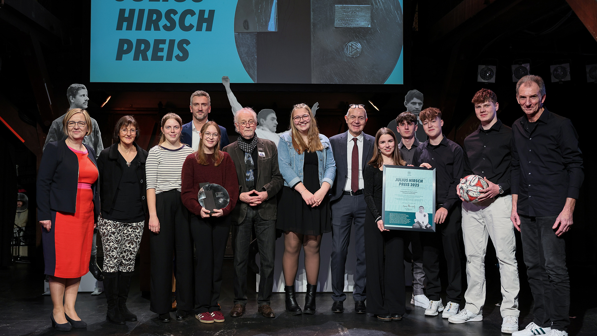 Eine Gruppe von Menschen steht auf einer Bühne vor einer großen Leinwand mit der Aufschrift 'JULIUS HIRSCH PREIS'. Einige Personen halten Auszeichnungen: eine Person hält eine runde Trophäe, eine andere eine Urkunde, und eine weitere Person hält einen Fußball. Die Personen tragen unterschiedliche Kleidung, darunter ein rotes Kleid, ein gestreiftes Oberteil, ein Anzug mit Krawatte und legere Kleidung. Die Bühne ist dunkel, und im Hintergrund sind weitere Auszeichnungen sichtbar.