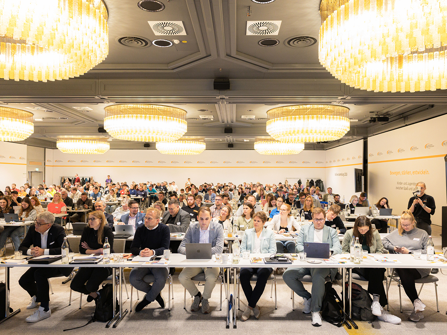 Eine große Gruppe von Menschen sitzt in einem Konferenzraum, viele arbeiten an Laptops. Über dem Publikum hängen mehrere große, runde Kronleuchter. Die Atmosphäre ist geschäftig und konzentriert, mit Tischen, die mit Wasserflaschen und Notizen ausgestattet sind.