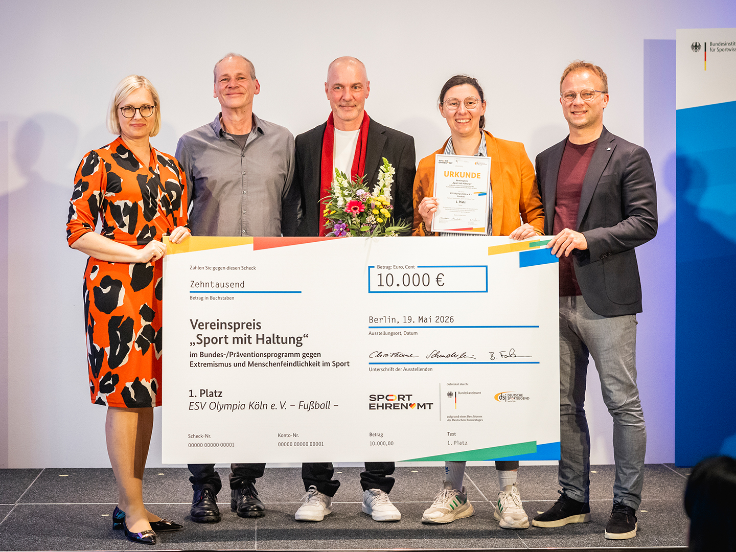 Eine Gruppe von fünf Personen steht auf einer Bühne und hält einen großen Scheck über 10.000 Euro. Die Personen sind in formeller Kleidung und lächeln. Im Hintergrund sind Banner und eine Präsentationsfläche sichtbar. Der Scheck ist mit "Vereinspreis Sport mit Haltung" beschriftet.
