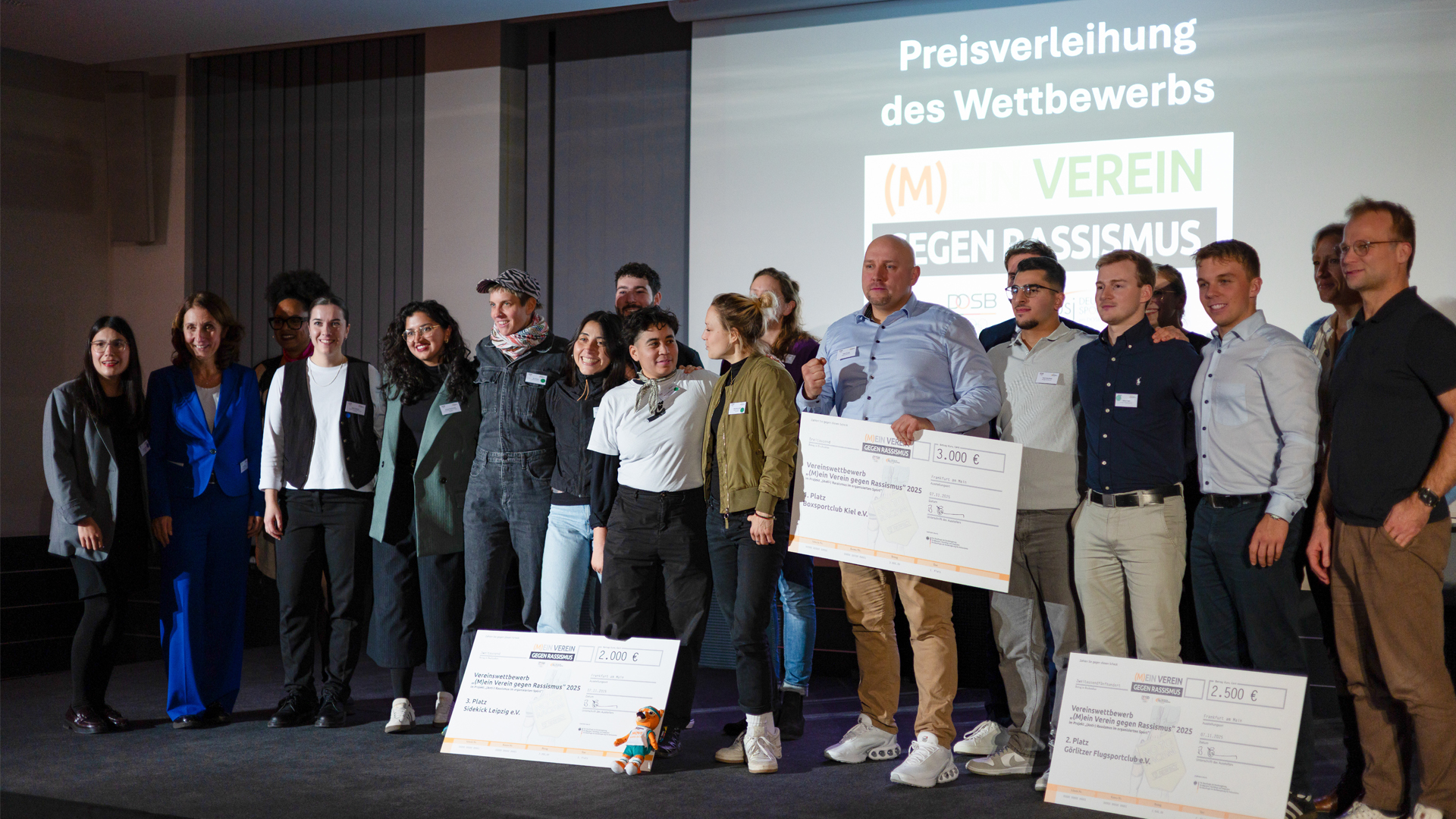 Eine Gruppe von Personen steht nebeneinander auf einer Bühne vor einer Leinwand mit der Projektion 'Preisverleihung des Wettbewerbs (M) Verein gegen Rassismus'. Einige Personen halten große symbolische Schecks in den Händen. Die Personen tragen unterschiedliche Kleidung, darunter Anzüge, Hemden und Freizeitkleidung. Die Szene wirkt formell und feierlich, passend zu einer Auszeichnung oder Preisverleihung im sportlichen Kontext.