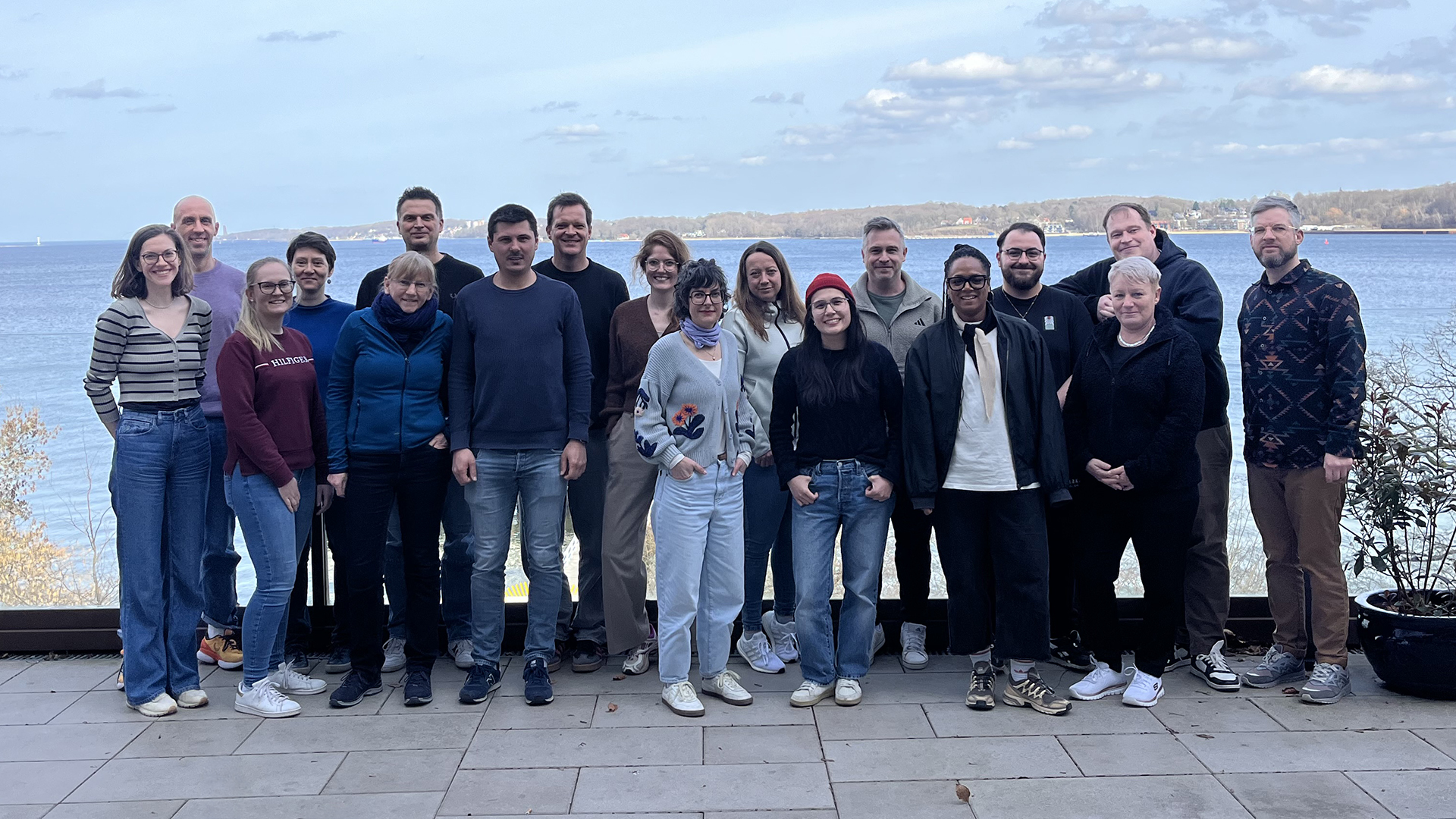 Eine Gruppe von Teilnehmenden des Vernetzungstreffens Z:T steht nebeneinander auf einer Terrasse am Wasser, im Hintergrund sind See und bewaldetes Ufer zu sehen.