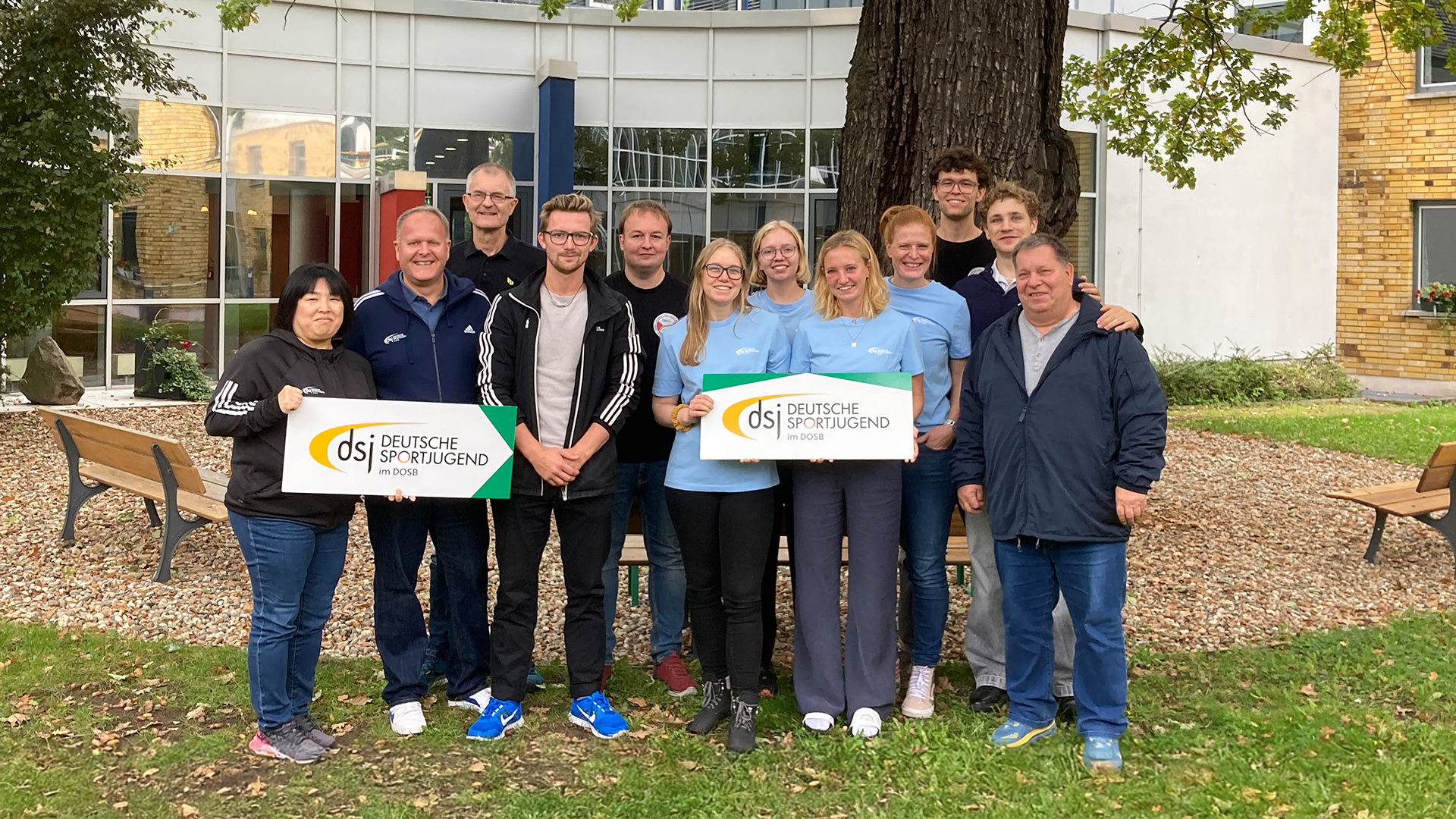 Gruppe von Personen steht im Freien vor einem Baum und hält zwei Schilder mit dem Logo der Deutschen Sportjugend, Vorbereitungstreffen dsj-Fachkräfteaustausch Japan 2025.