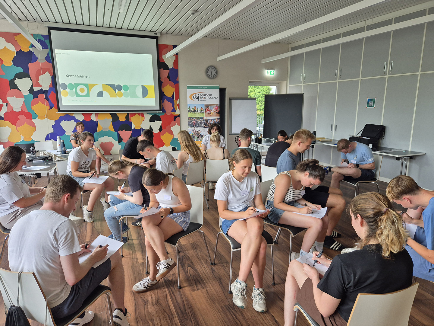 Eine Gruppe von jungen Menschen sitzt in einem Raum und schreibt auf Papier. Im Hintergrund ist eine bunte Wand mit verschiedenen Gesichtern zu sehen. Ein Bildschirm zeigt eine Präsentation. Die Atmosphäre wirkt konzentriert und aktiv.