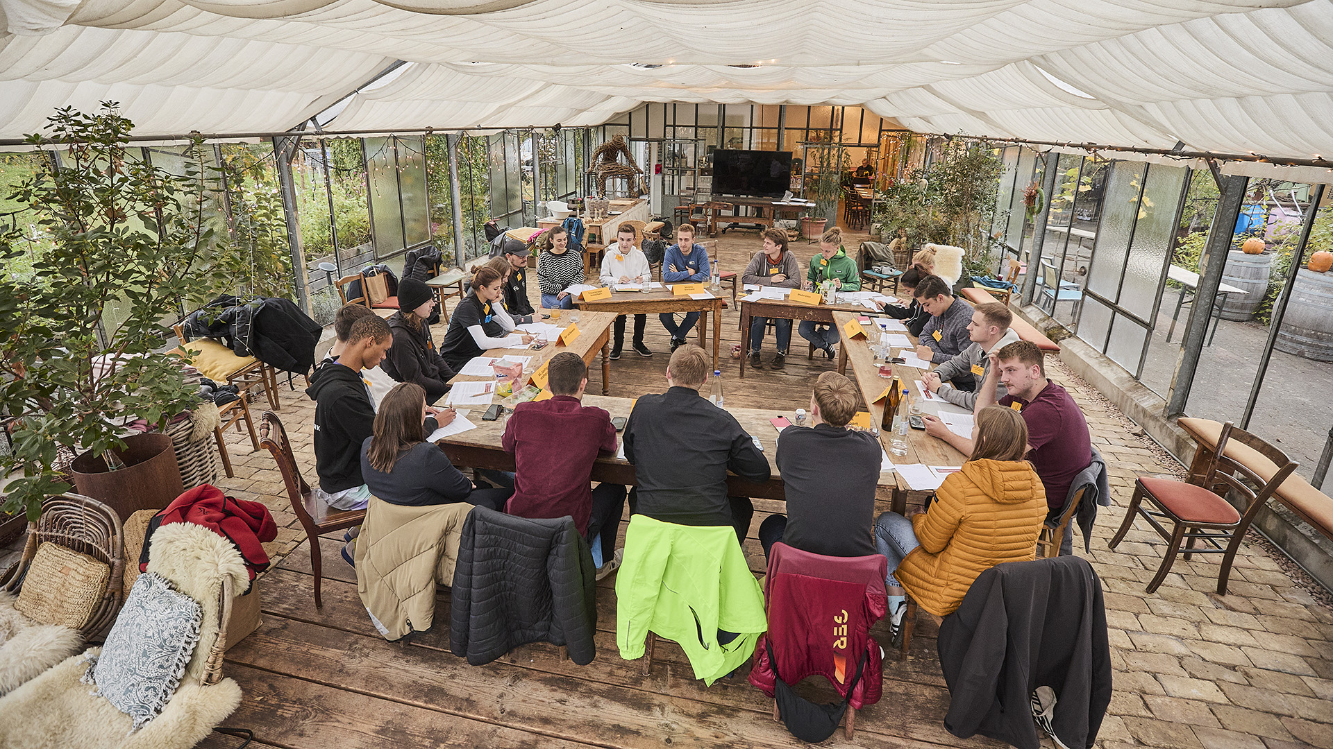 In einem hellen, mit Pflanzen dekorierten Gewächshaus sitzen junge Menschen an einem großen, hufeisenförmigen Holztisch. Sie sind in eine Diskussion vertieft und haben verschiedene Unterlagen, Stifte und Getränke vor sich liegen. Die Kleidung ist bunt und lässig, darunter Jacken und Pullover. Im Hintergrund steht ein großer Fernseher und weitere Sitzgelegenheiten sind entlang der Glaswände verteilt. Die Atmosphäre wirkt offen und gemeinschaftlich, passend zu einer Veranstaltung, bei der junge Engagierte im organisierten Sport zusammenkommen.