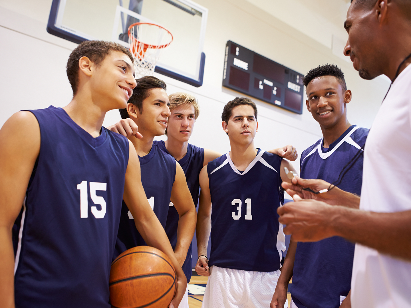 Eine Gruppe von fünf Basketballspielern in blauen Trikots steht zusammen in einer Sporthalle. Sie hören einem Trainer zu, der ihnen etwas erklärt. Ein Spieler hält einen Basketball in der Hand.