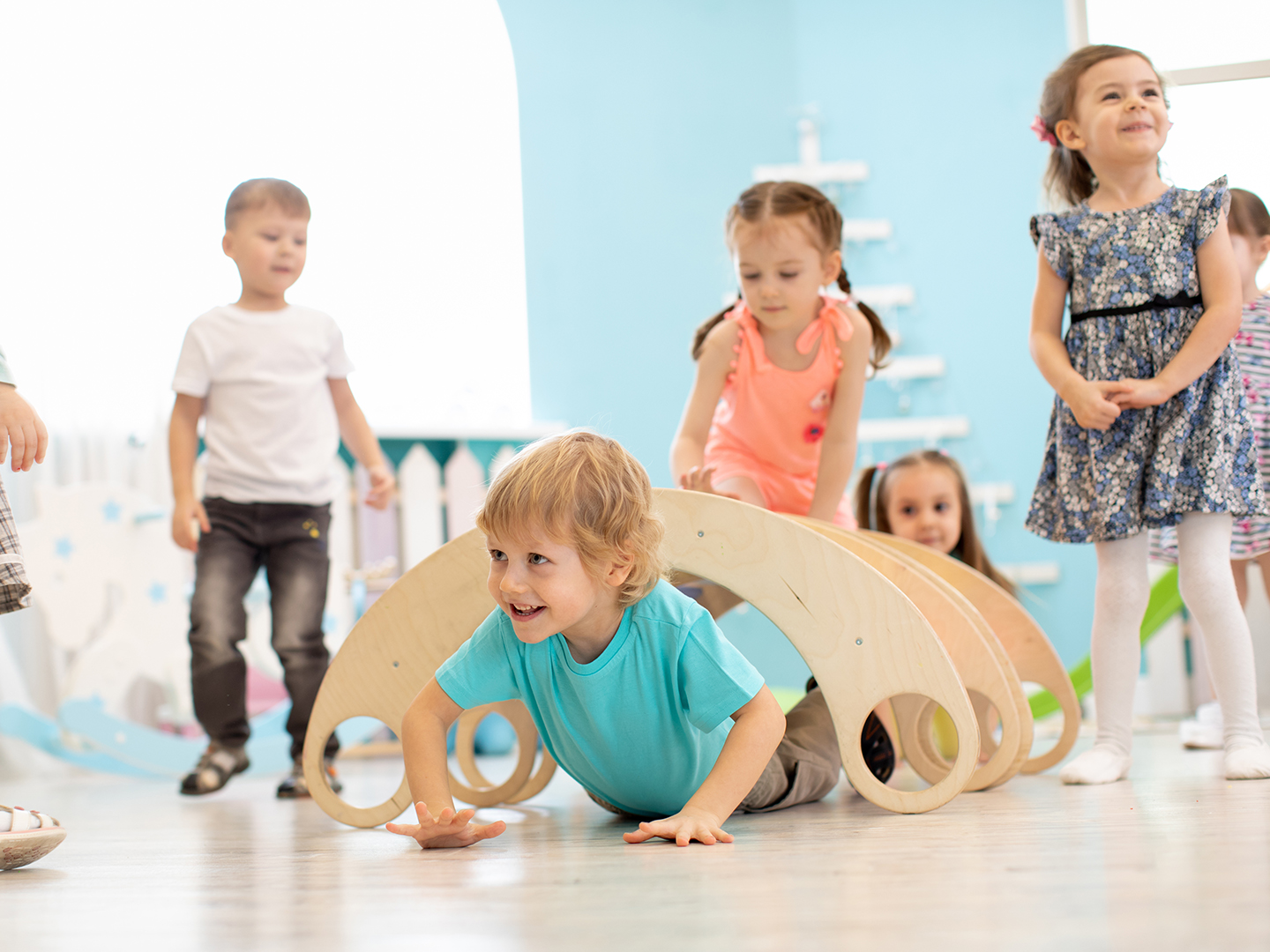 Eine Gruppe von Kindern spielt in einem hellen Raum. Ein Junge krabbelt unter einer hölzernen Spielstruktur hindurch, während andere Kinder in der Nähe stehen und zuschauen. Einige Kinder lachen und haben Spaß.