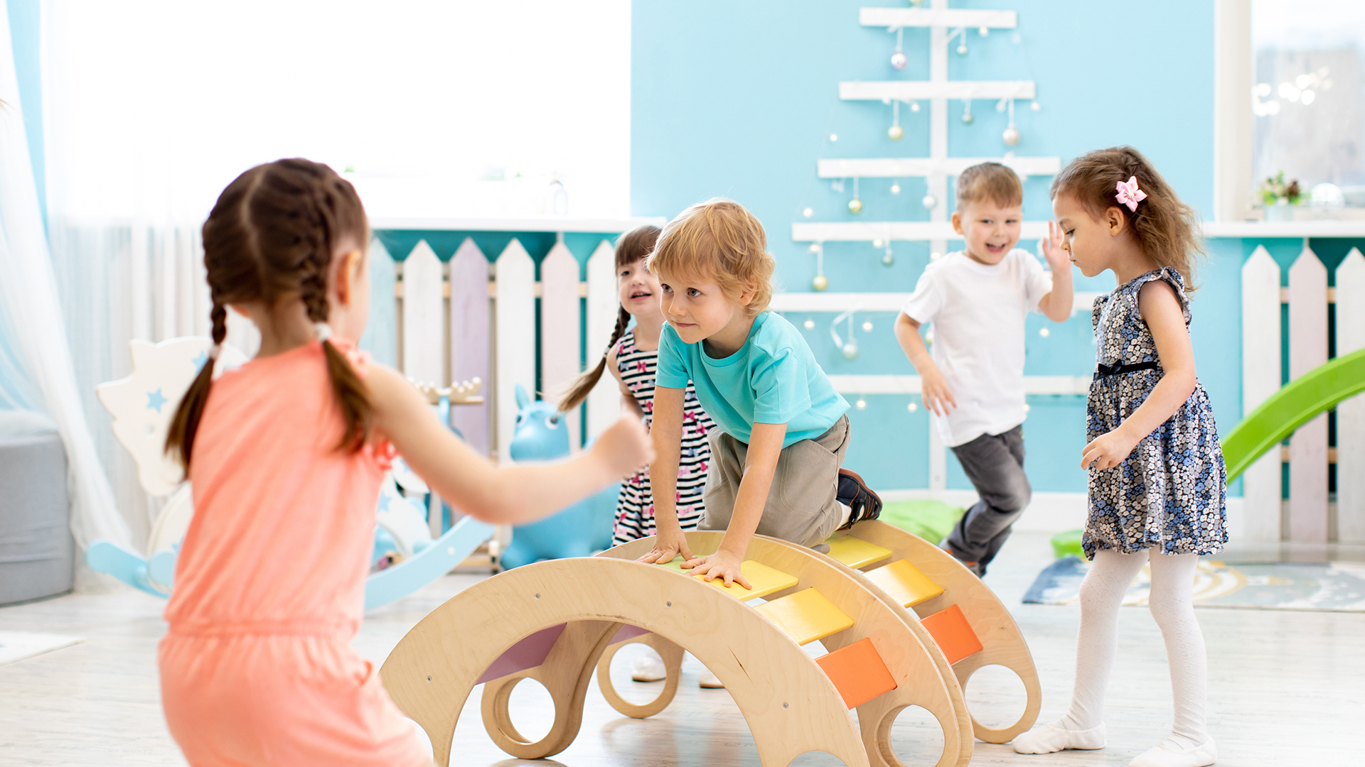 Mehrere Kinder spielen und bewegen sich gemeinsam in einem hellen Bewegungsraum, klettern auf bunten Holz‑Spielgeräten und fördern dabei Gleichgewicht, Koordination und Freude an Bewegung.