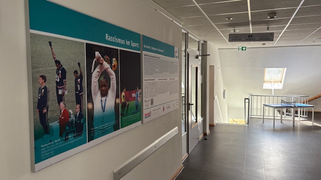 Wand mit zwei großen Plakaten zur Demokratieausstellung in Jugendbildungsstätten. Links ein Bild von Sportlern, rechts Text über Rassismus im Sport