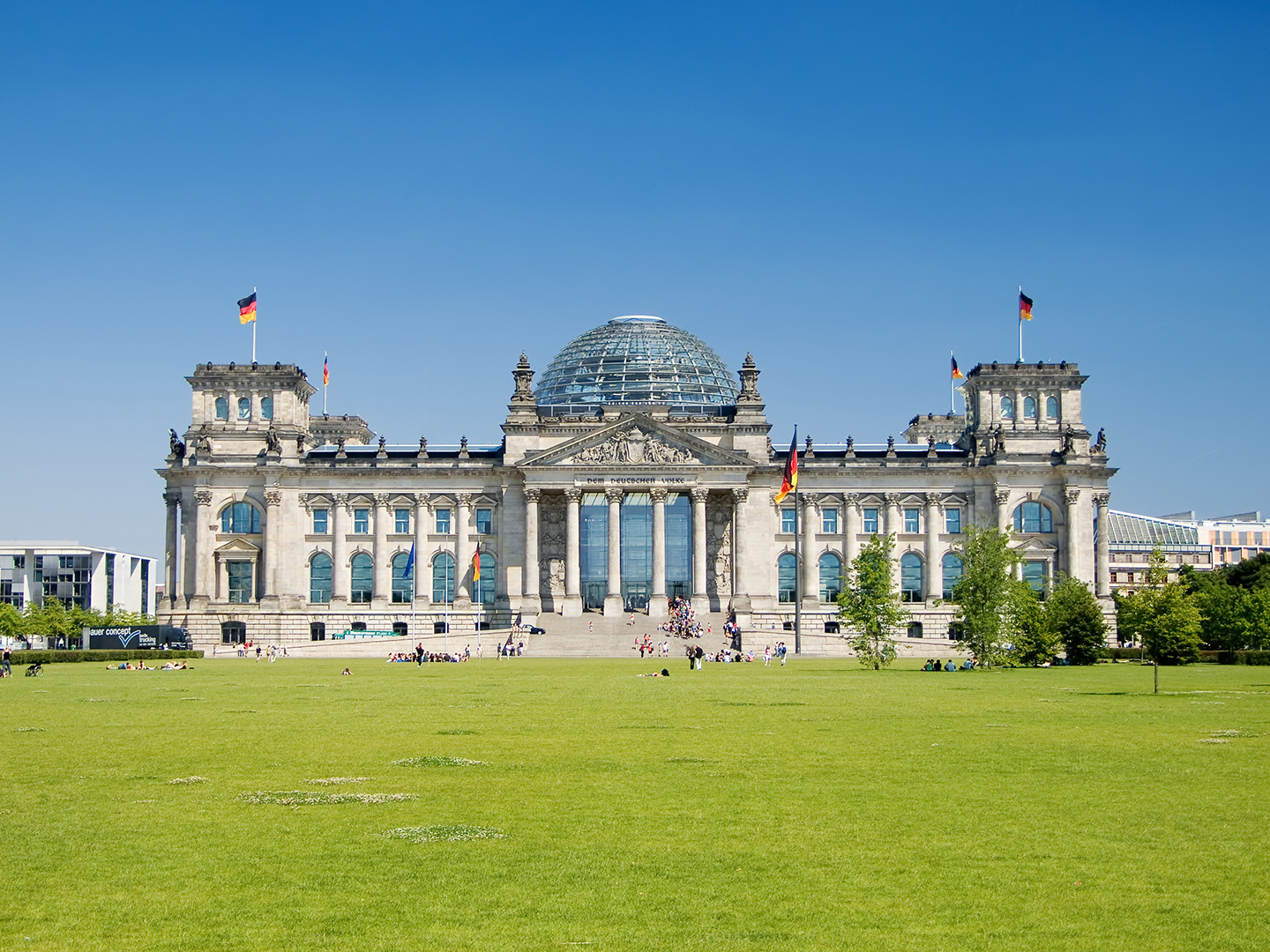 Das Bild zeigt das Reichstagsgebäude in Berlin an einem sonnigen Tag. Die beeindruckende Fassade mit der Glaskuppel ist sichtbar, umgeben von einer grünen Wiese. Einige Menschen sind im Vordergrund zu sehen, und die deutschen Flaggen wehen an den Dächern.