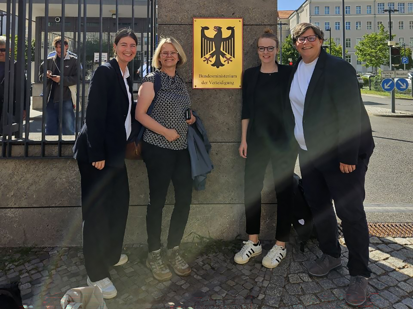 Vier Personen stehen lächelnd vor einem Schild des Bundesministeriums der Verteidigung in Deutschland. Sie tragen legere Kleidung und scheinen eine positive Atmosphäre zu haben. Im Hintergrund sind Bäume und ein Gebäude sichtbar.