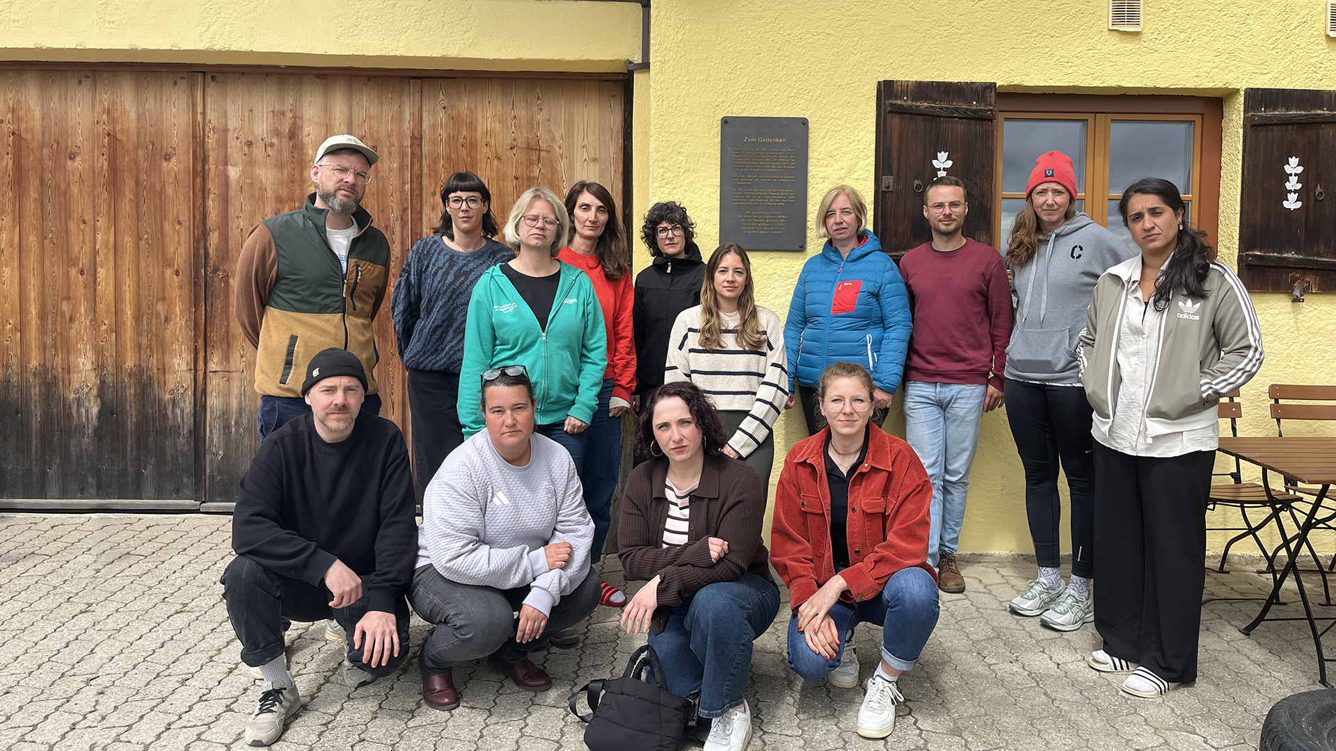 Gruppe von Teilnehmenden einer Erinnerungsfahrt der Deutschen Sportjugend steht und kniet vor einem Gebäude am Sudelfeld; im Hintergrund ist eine Gedenktafel zur Gründung angebracht.