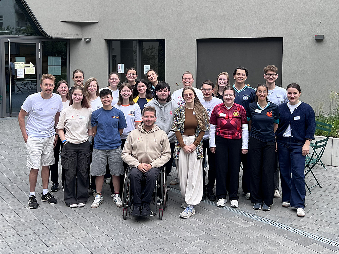 Eine Gruppe von 22 Personen steht zusammen im Freien vor einem Gebäude. Eine Person sitzt im Rollstuhl. Die Gruppe lächelt und posiert für ein Foto. Die Personen sind eher sportlich gekleidet.