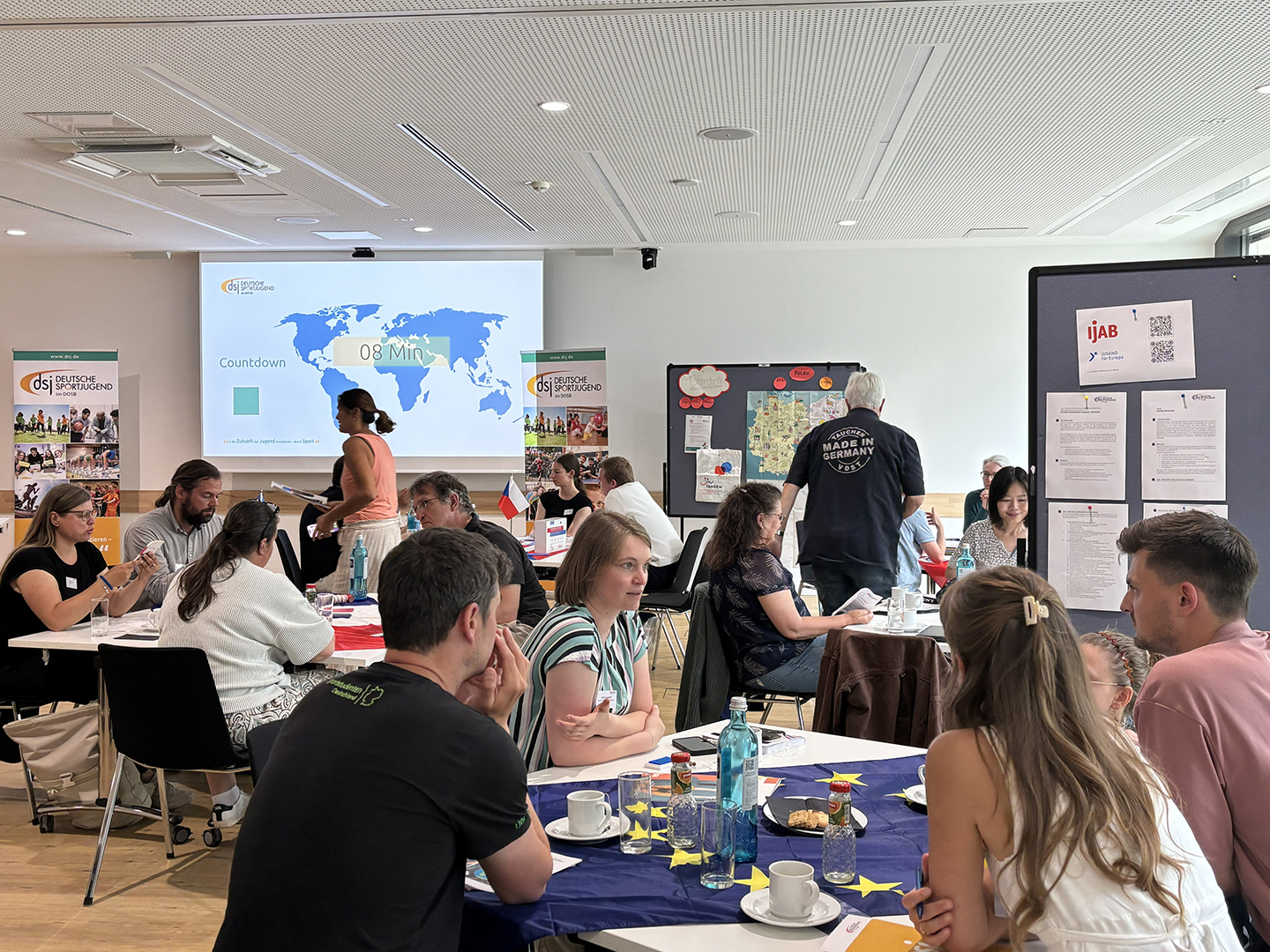 Mehrere Gruppen von Menschen sitzen an Tischen in einem Raum, während eine Präsentation mit einer Weltkarte im Hintergrund gezeigt wird. Einige Teilnehmer diskutieren, während andere Notizen machen oder auf den Bildschirm schauen.