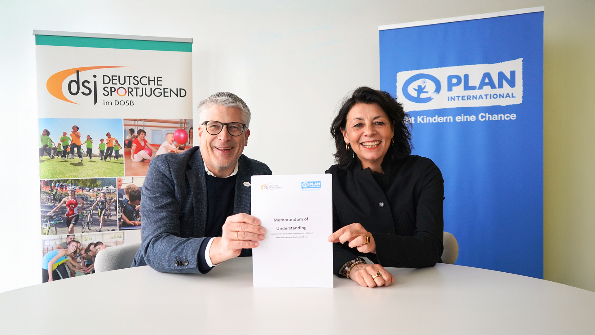 Die Deutsche Sportjugend und Plan International unterzeichnen ein Memorandum of Understanding zur Stärkung von Kinderrechten im Sport. Zwei Vertreter*innen der Organisationen sitzen an einem Tisch und halten das unterschriebene Dokument vor Bannern beider Institutionen.