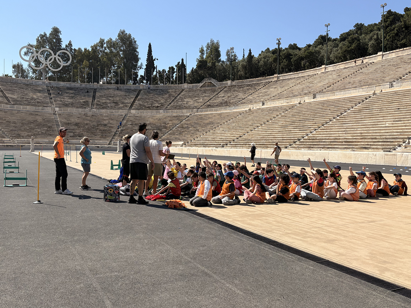 Eine Gruppe von Kindern in orangefarbenen T-Shirts sitzt auf dem Boden eines Stadions. Einige Erwachsene stehen vor ihnen und erklären etwas. Im Hintergrund sind die Olympischen Ringe und leere Sitzreihen zu sehen.