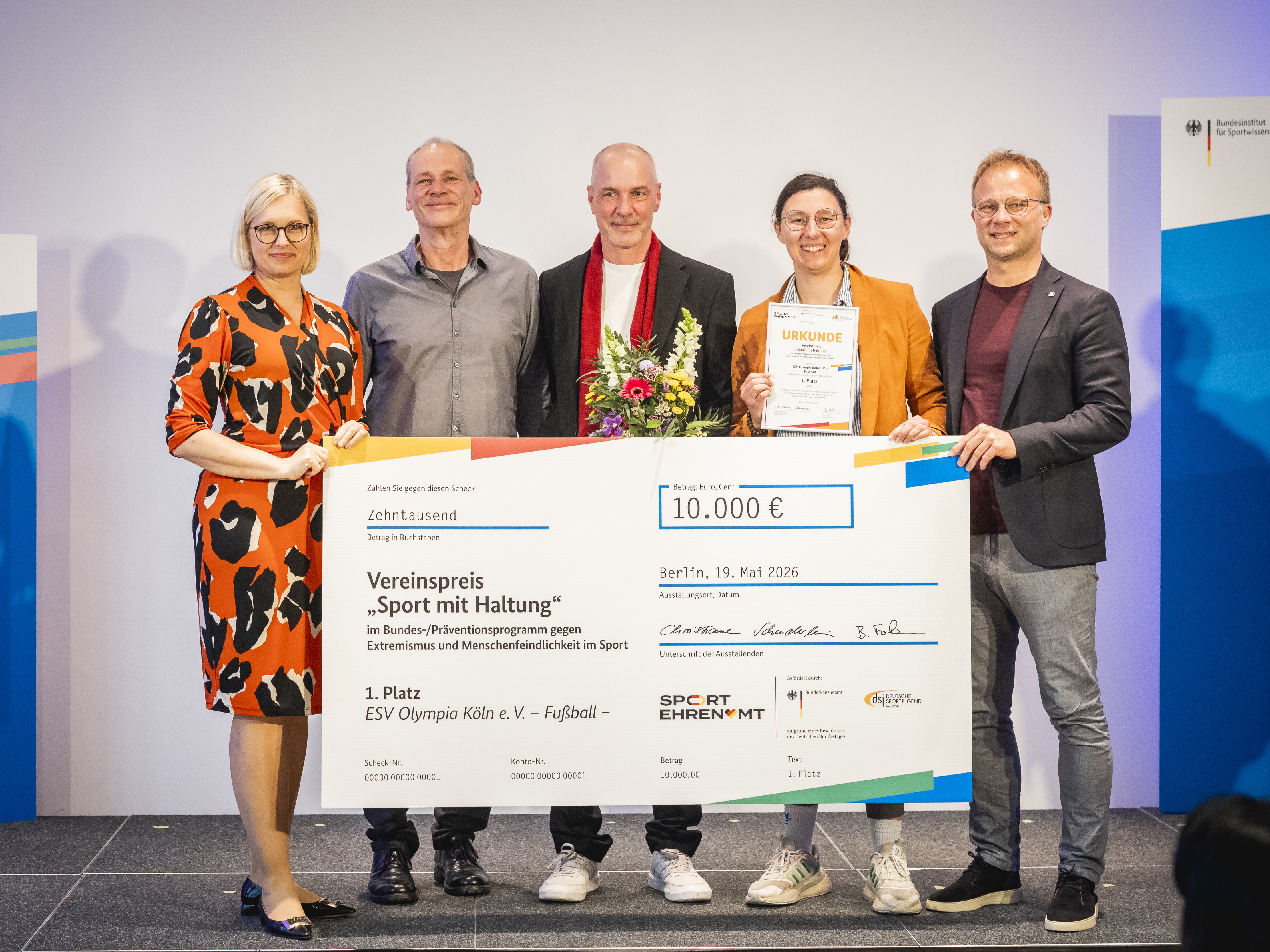 Eine Gruppe von fünf Personen steht auf einer Bühne und hält einen großen Scheck über 10.000 Euro. Der Scheck ist für den Vereinspreis "Sport mit Haltung" und wird an ESV Olympia Köln e.V. für ihre Leistungen im Sport verliehen.