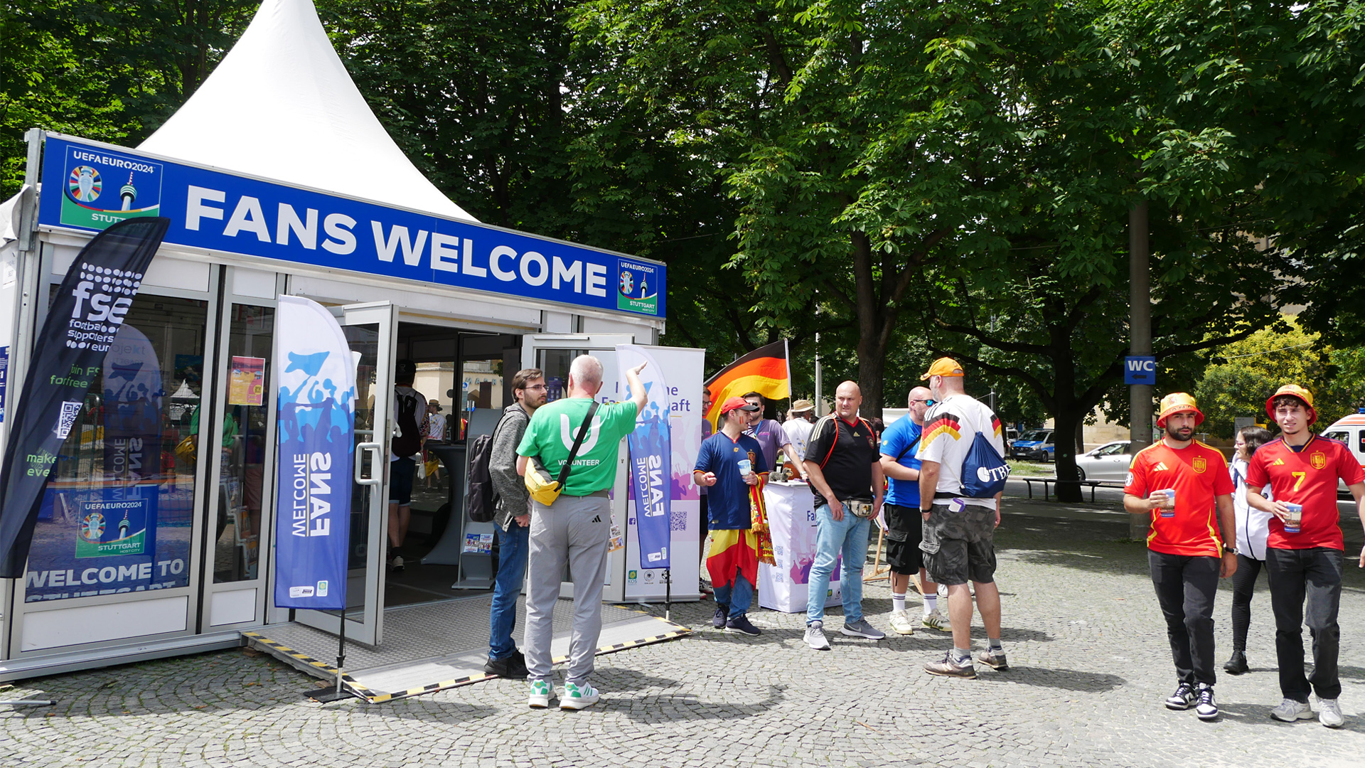 Zelt mit Schriftzug 'FANS WELCOME' auf einem Fanprojekt-Event, mehrere Personen in Fußballtrikots und mit Deutschlandfahnen stehen davor