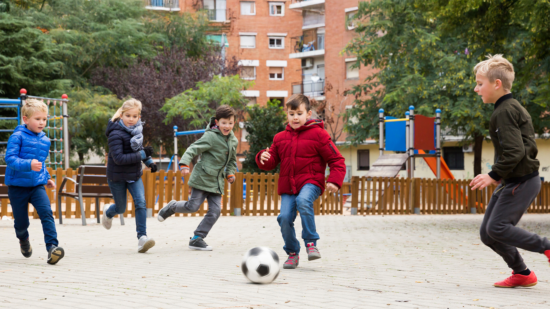 Fünf Kinder spielen Fußball auf einem Schulhof mit Klettergerüst und Wohngebäuden im Hintergrund.