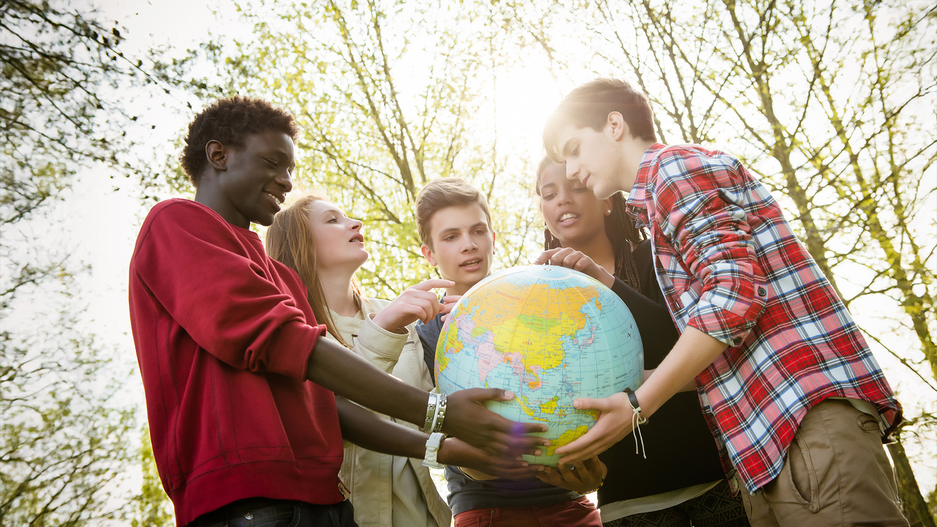 Sechs Jugendliche halten gemeinsam einen Globus, umgeben von Bäumen. Der Globus zeigt eine Weltkarte mit Kontinenten und Ländern. Fortbildung Sport kennt keine Grenzen für internationale Jugendbegegnungen im Sport.