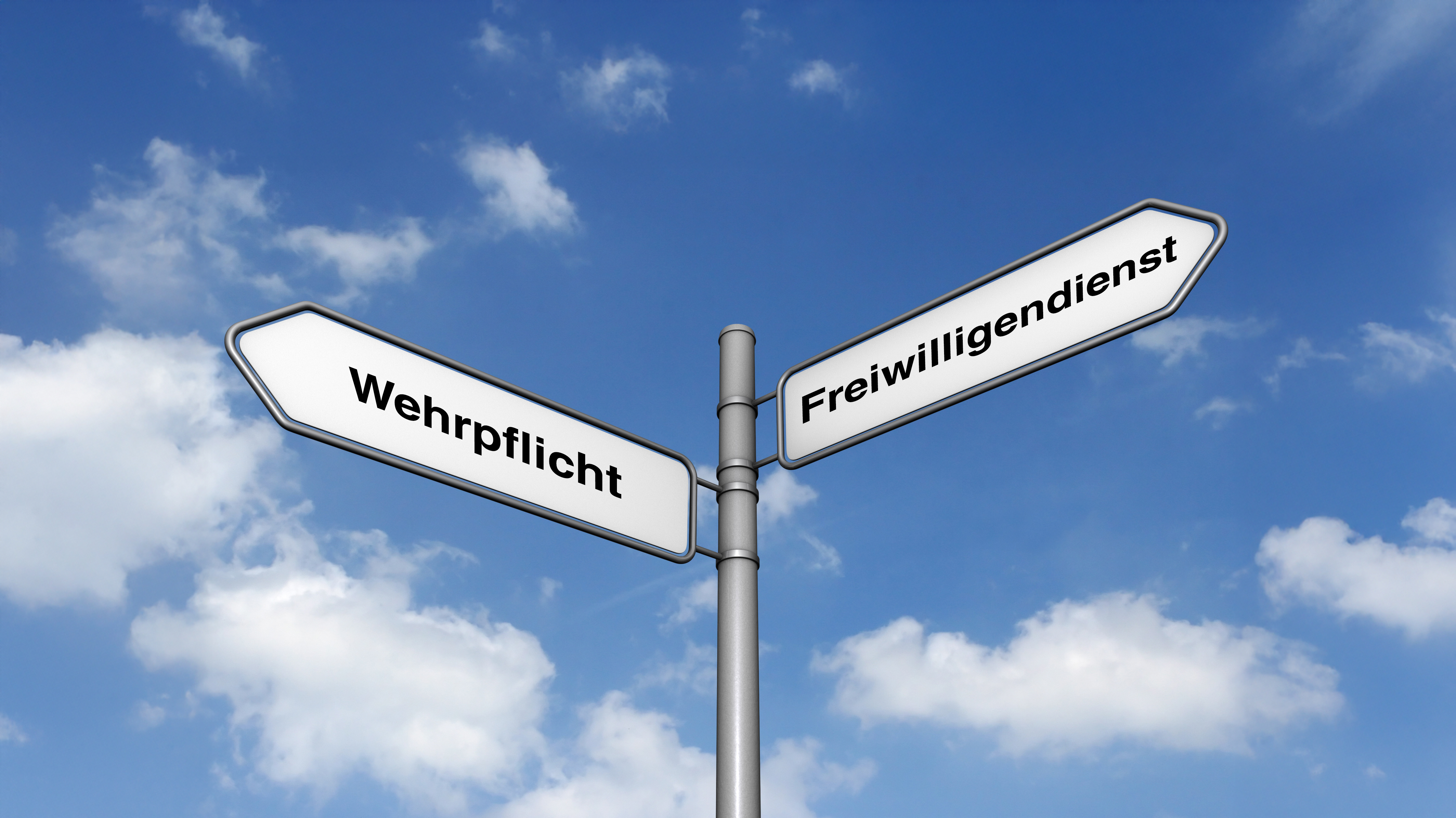 Ein metallener Wegweiser mit zwei weißen, rechteckigen Schildern, die jeweils an einem Ende spitz zulaufen. Das linke Schild zeigt nach links und trägt die Aufschrift 'Wehrpflicht'. Das rechte Schild zeigt nach rechts und trägt die Aufschrift 'Freiwilligendienst'. Der Wegweiser steht vor einem blauen Himmel mit einigen weißen Wolken.