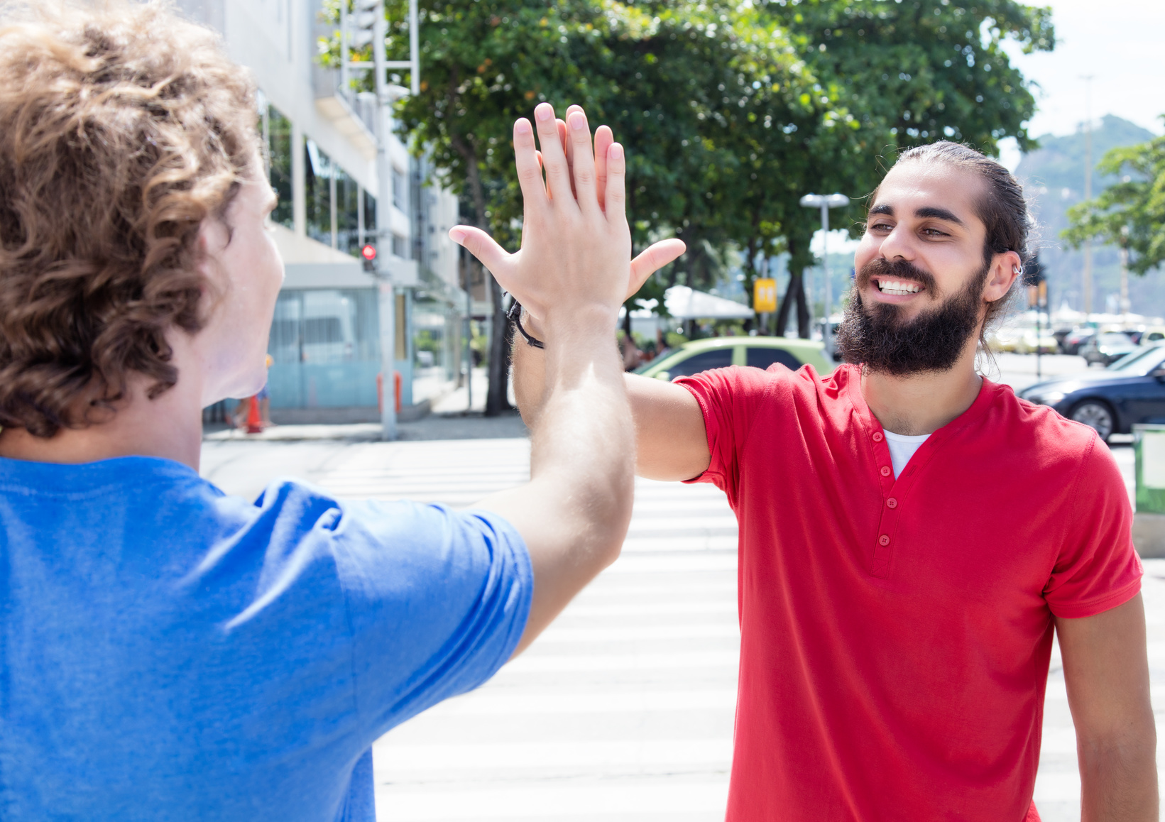 Zwei Männer stehen sich gegenüber und geben sich ein High-Five. Der Mann links trägt ein blaues T-Shirt und hat lockige Haare, während der Mann rechts ein rotes T-Shirt trägt und einen Bart hat. Im Hintergrund sind Bäume und parkende Autos sichtbar.