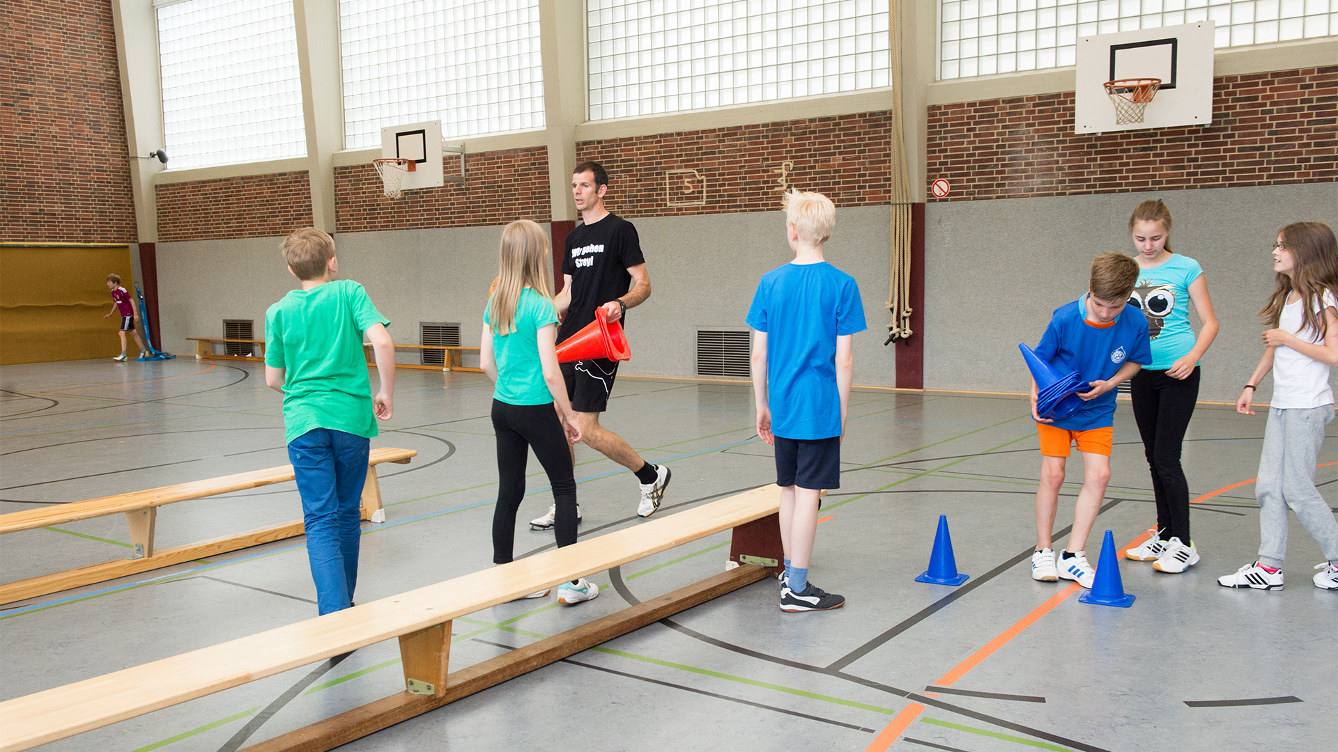Sechs Kinder in Sportkleidung stehen in einer Turnhalle. Ein Erwachsener trägt ein schwarzes T-Shirt und läuft mit roten Objekten. Bänke und Hütchen sind sichtbar.