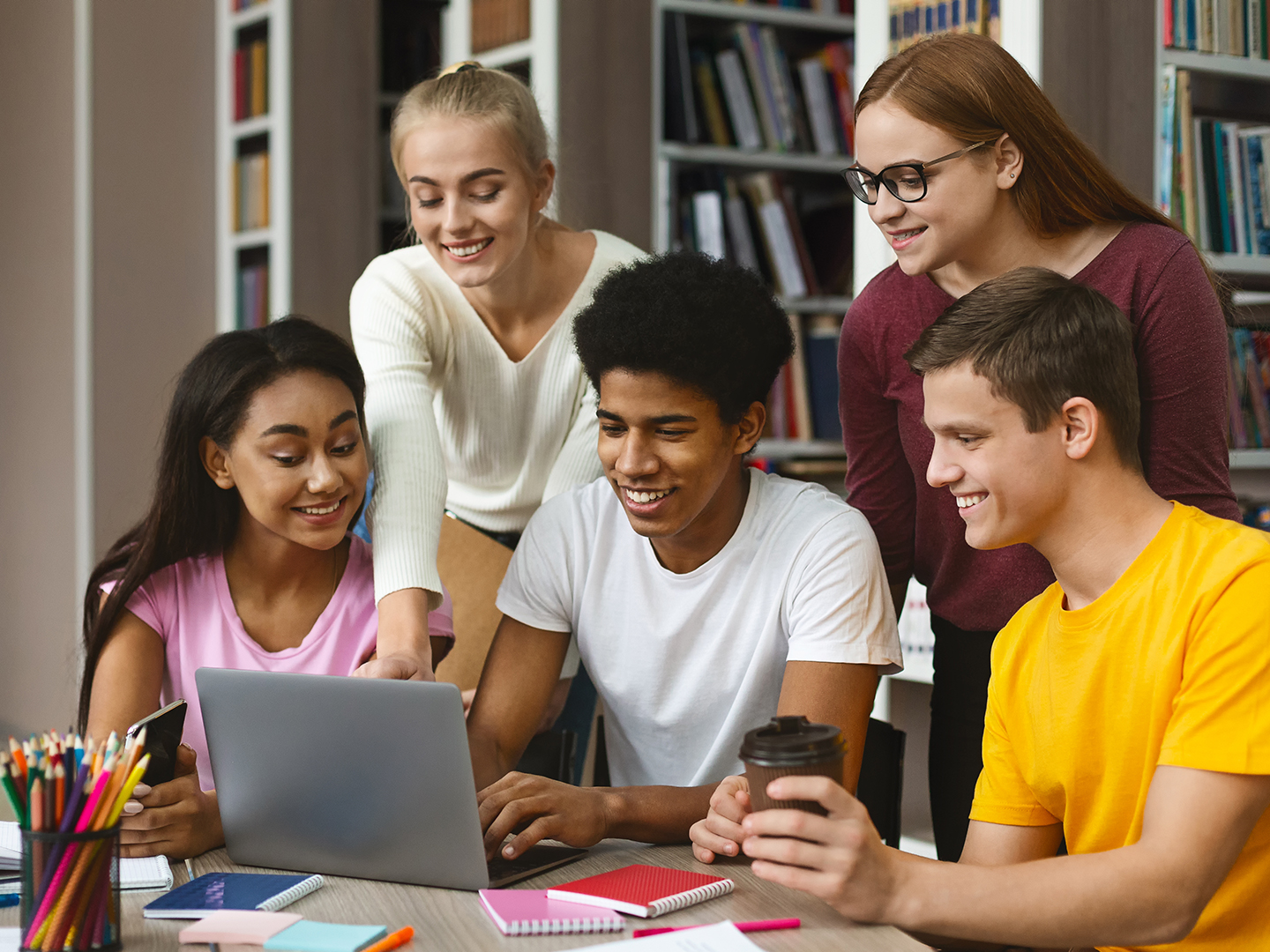 Fünf junge Menschen sitzen an einem Tisch in einer Bibliothek und schauen auf einen Laptop. Zwei Mädchen zeigen auf den Bildschirm, während die anderen aufmerksam zuschauen. Auf dem Tisch liegen Notizblöcke, Stifte und eine Kaffeetasse.